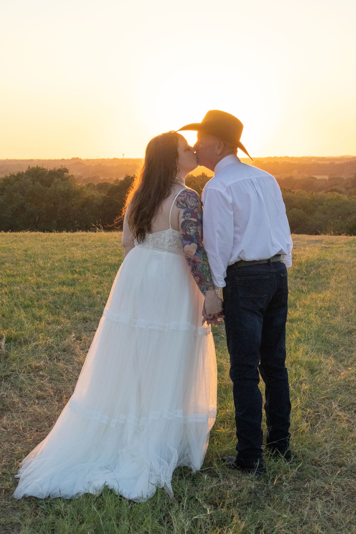 Couple sharing a romantic kiss at sunset in a green field, bride in white gown and groom in cowboy hat.