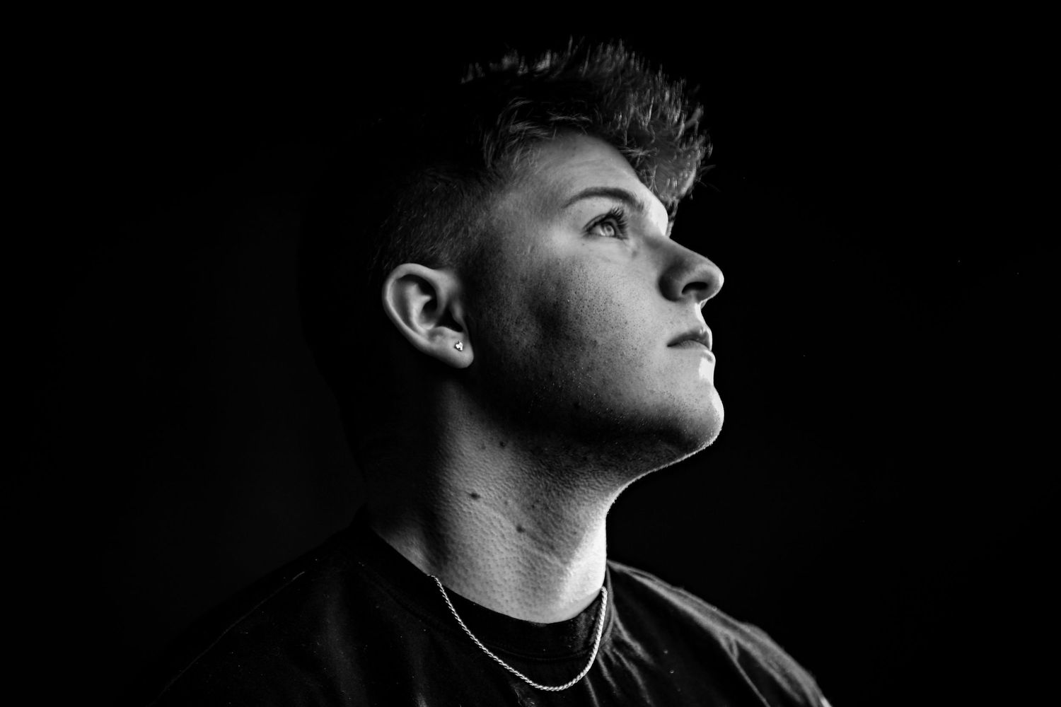 Black and white portrait of a young man in a black t-shirt looking upward against a dark background.