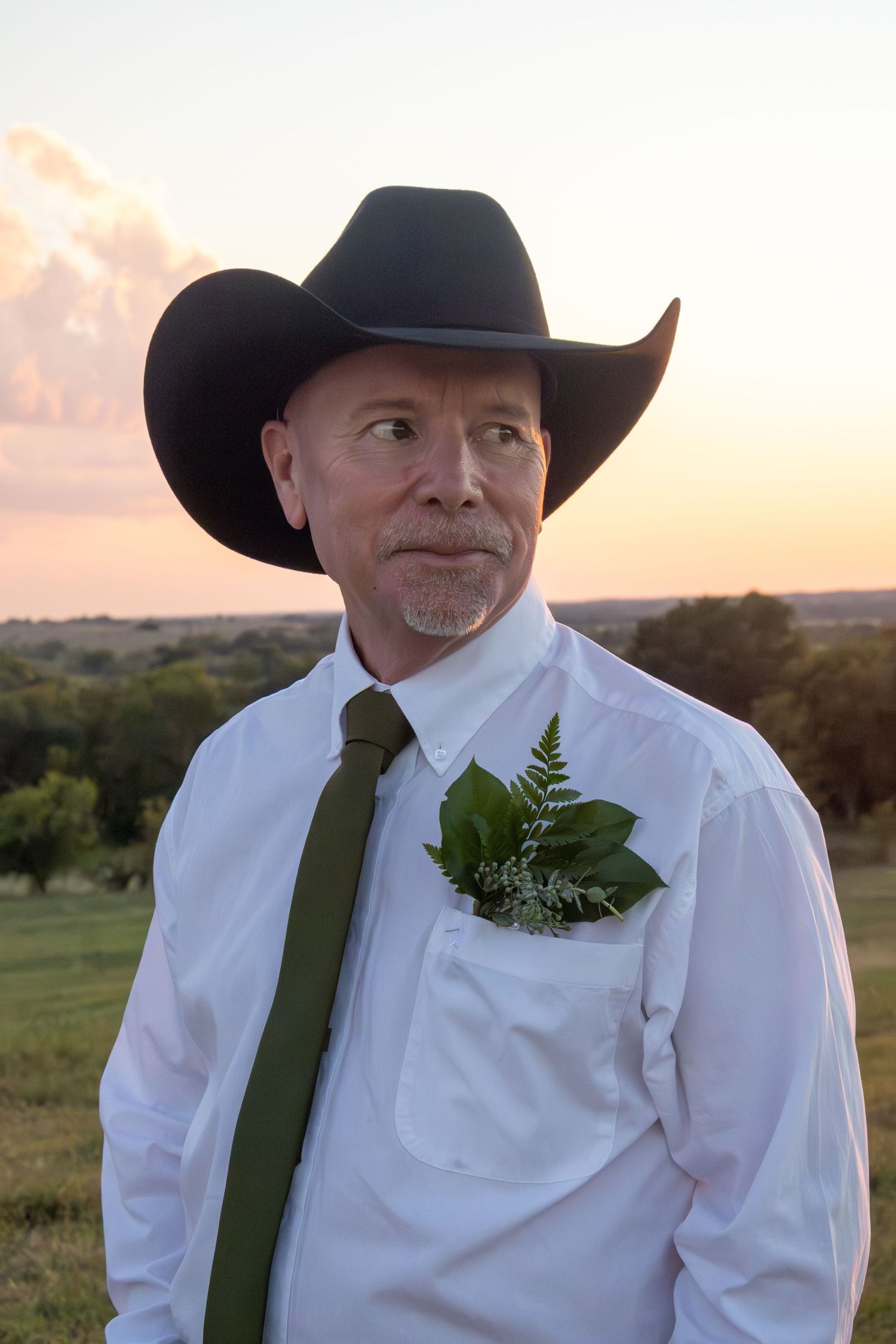 Groom in black cowboy hat, white shirt, and olive tie with boutonniere at sunset.
