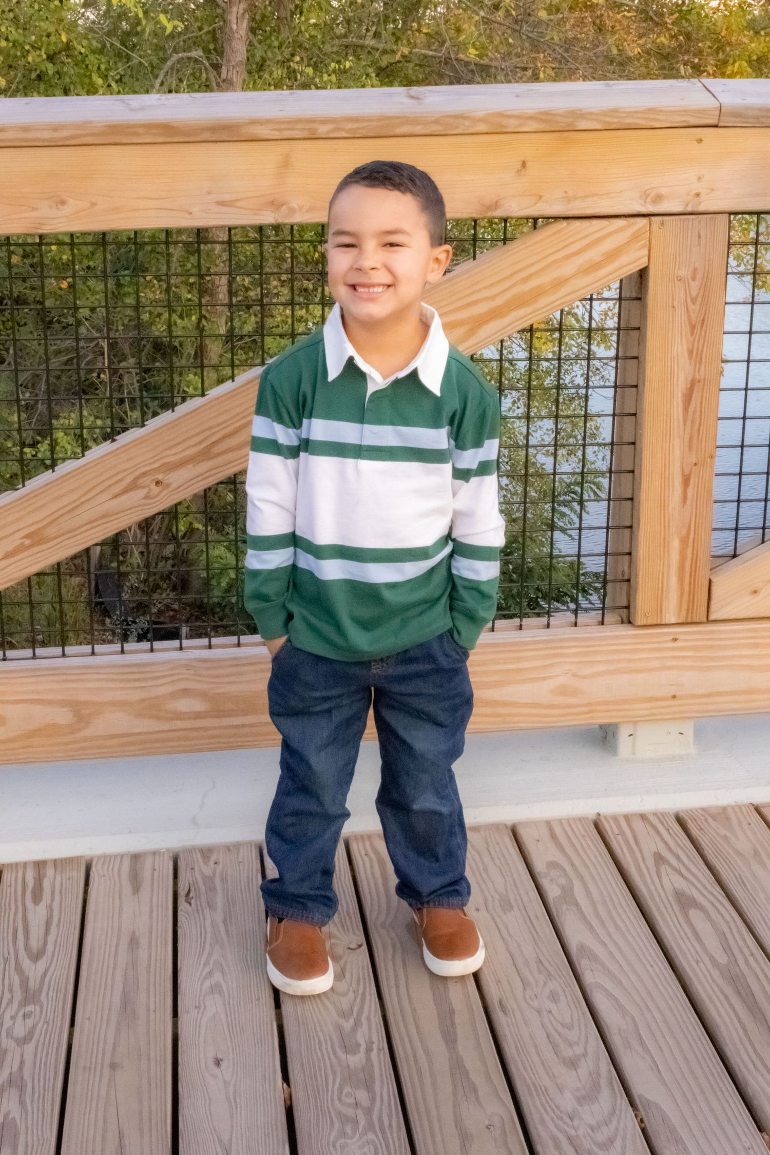 Smiling young boy in green striped polo and jeans standing on wooden bridge outdoors.