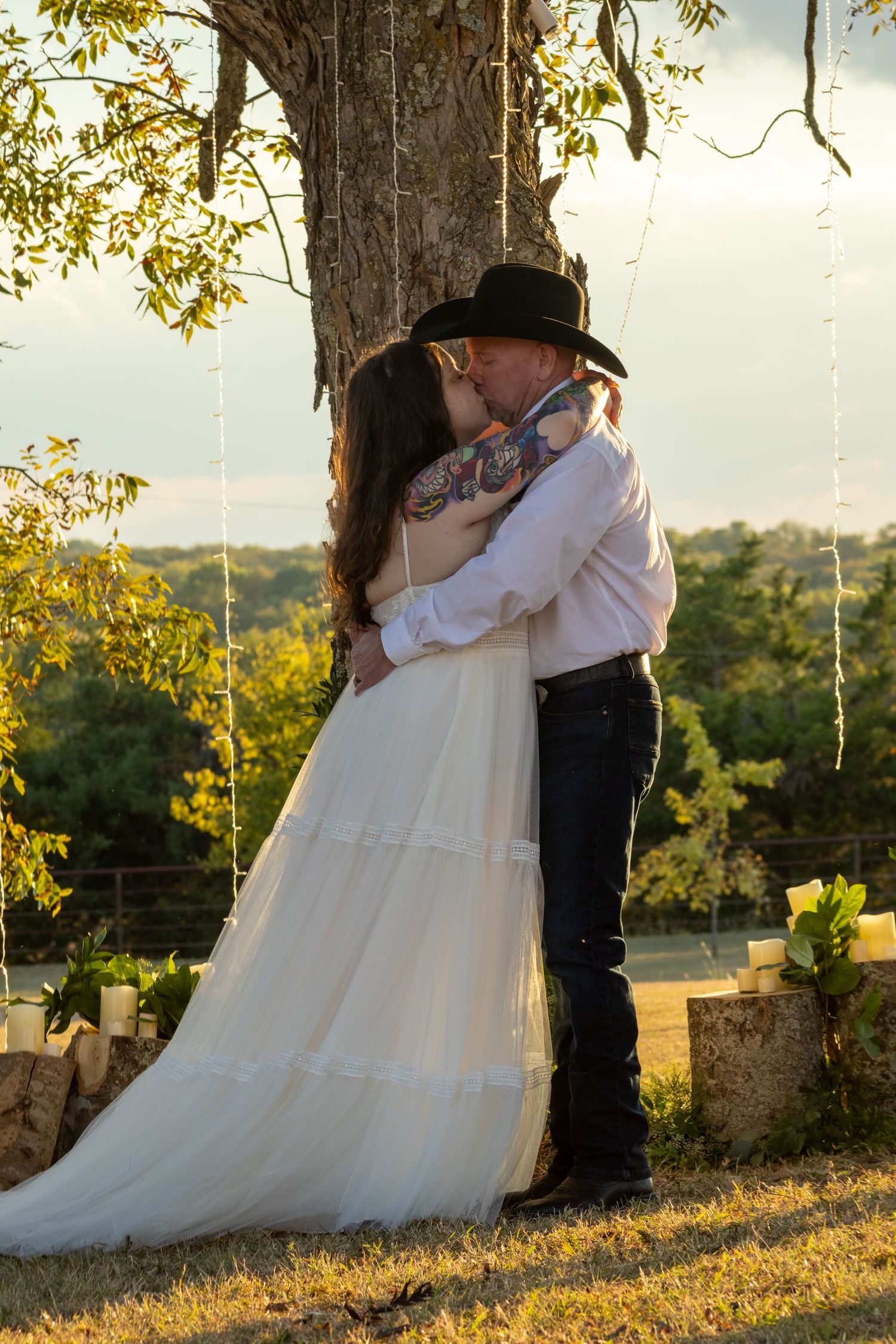 Couple sharing first kiss in wedding attire under tree at golden hour outdoor ceremony.