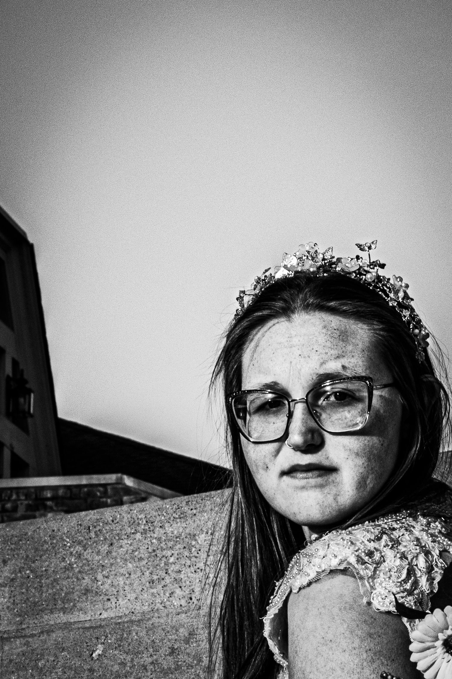 Black and white portrait of a young woman wearing a floral tiara and glasses, looking pensively at the camera outdoors.