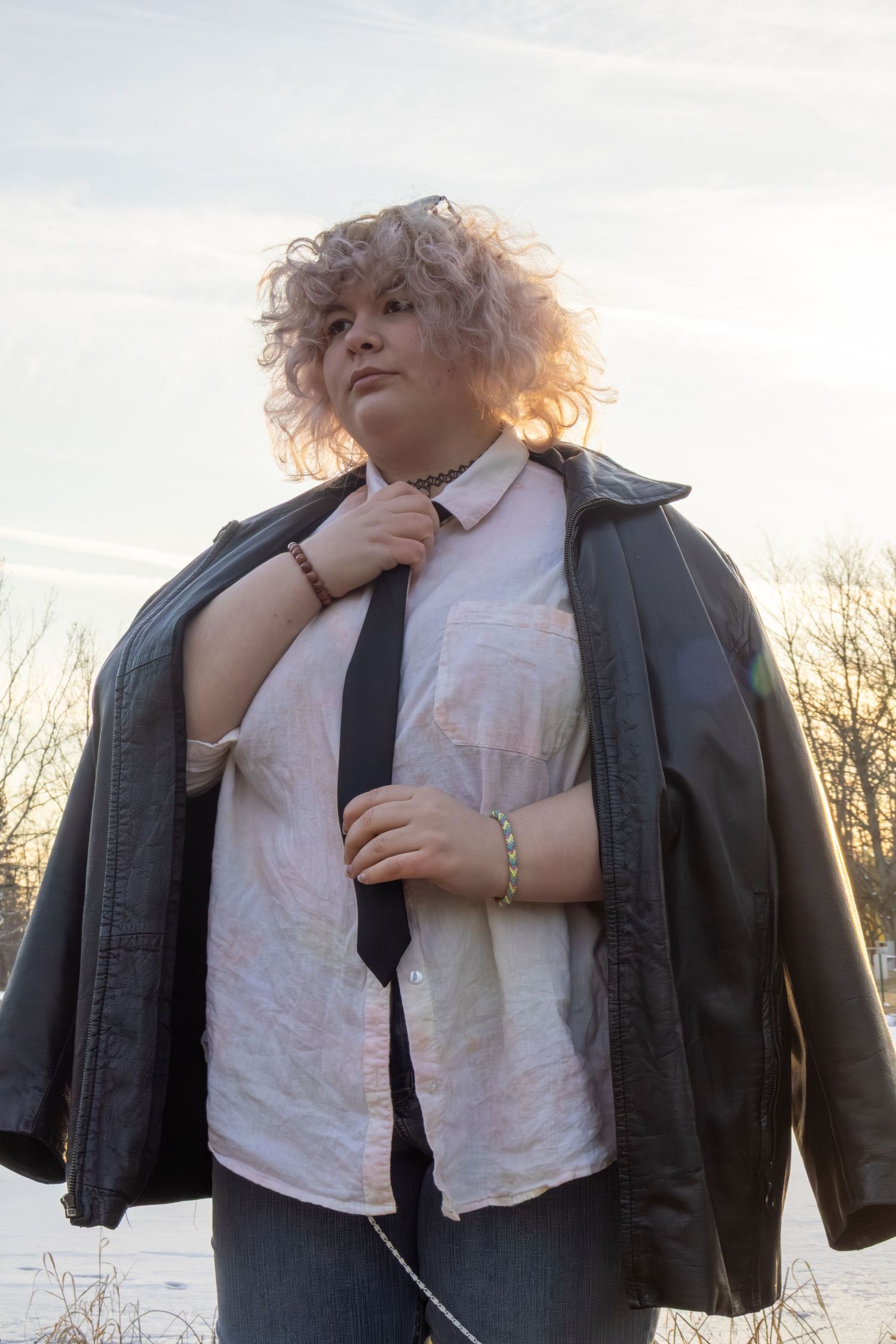 Woman with curly pink hair in leather jacket and tie posing outdoors in winter sunset.