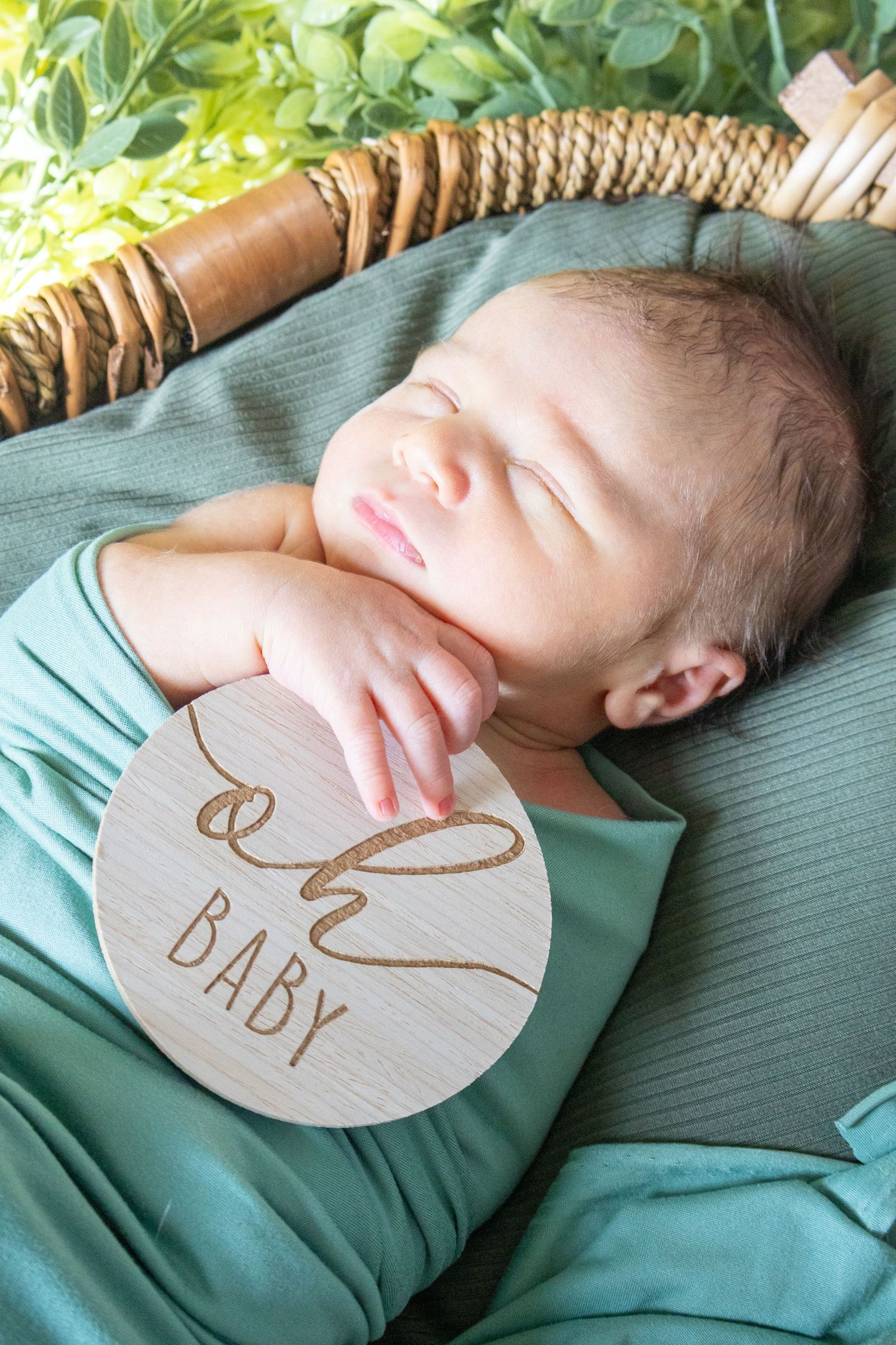 Sleeping newborn baby in green wrap holding wooden Oh Baby milestone disc in wicker basket.