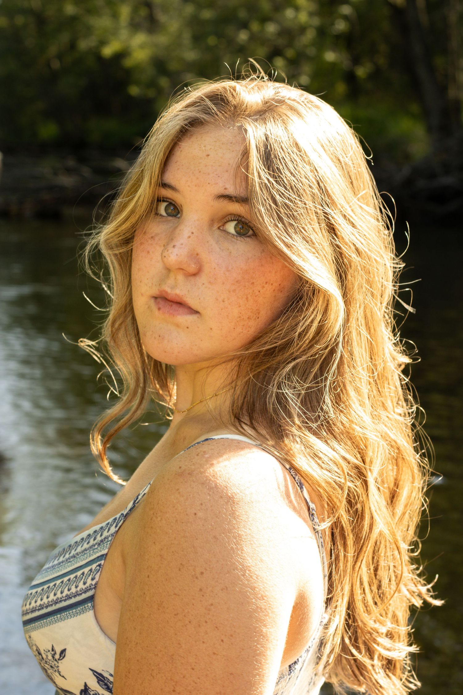 Young woman with freckles and blonde wavy hair posing near sunlit river in summer.