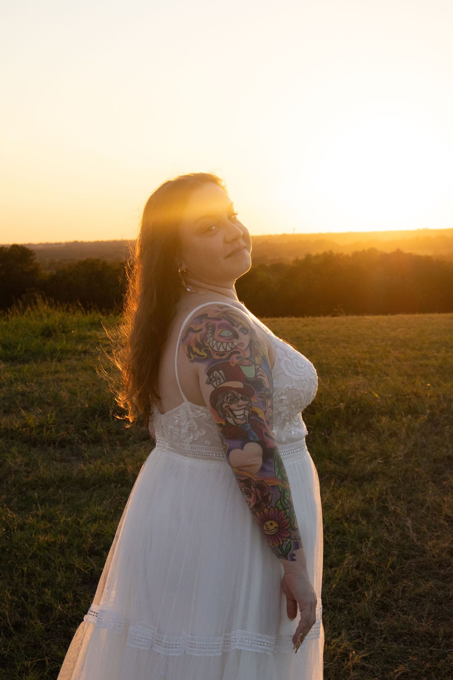 Bride in white dress with colorful sleeve tattoo posing at golden hour sunset outdoors.