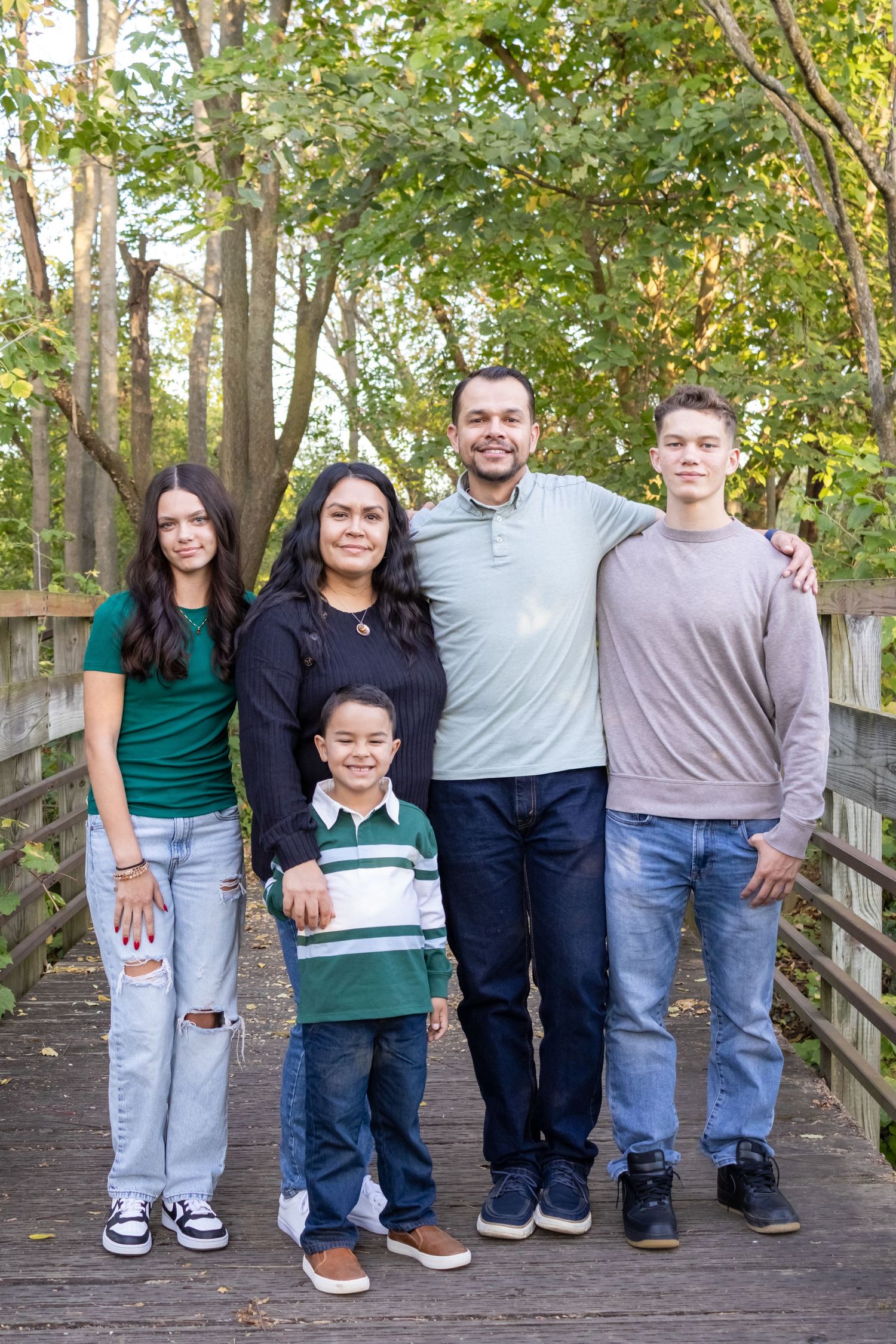 Happy family of five smiling together on wooden bridge surrounded by autumn trees in park.