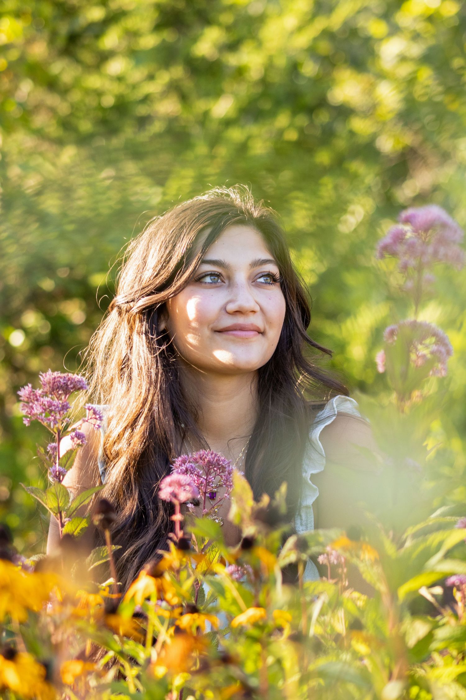 Smiling young woman with dark hair surrounded by colorful wildflowers in sunlit garden setting.