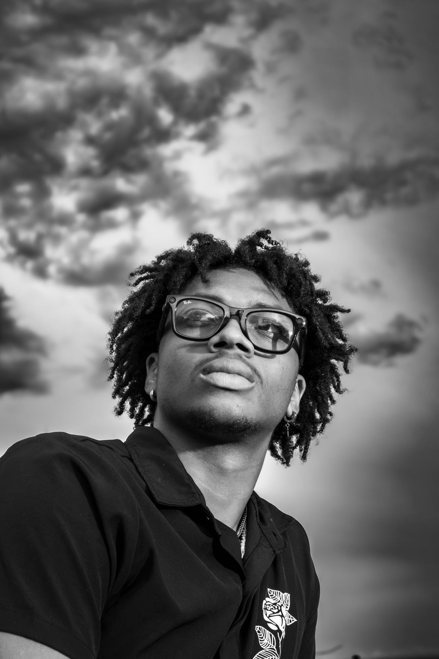 Black and white portrait of a young man with dreadlocks and glasses looking up against a dramatic cloudy sky.