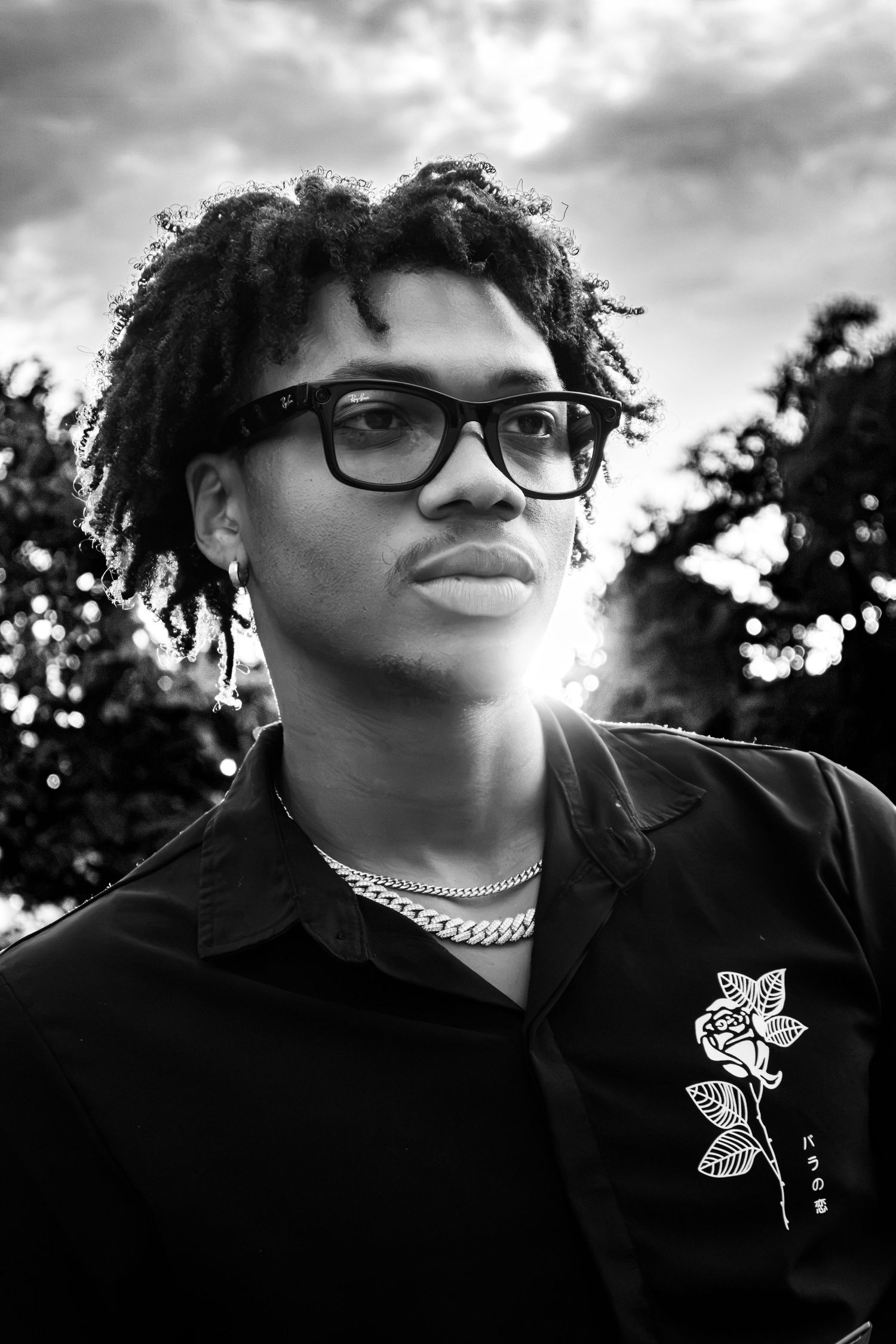 Black and white portrait of a young man wearing glasses, chain necklace, and black shirt with rose graphic outdoors.