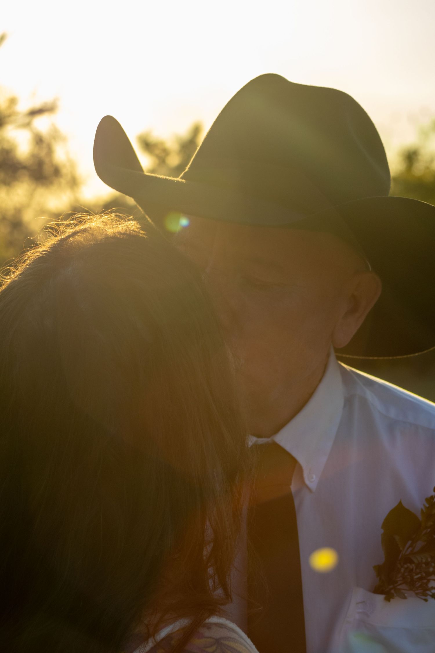 Couple sharing a romantic kiss at golden hour, groom wearing a cowboy hat and boutonniere.