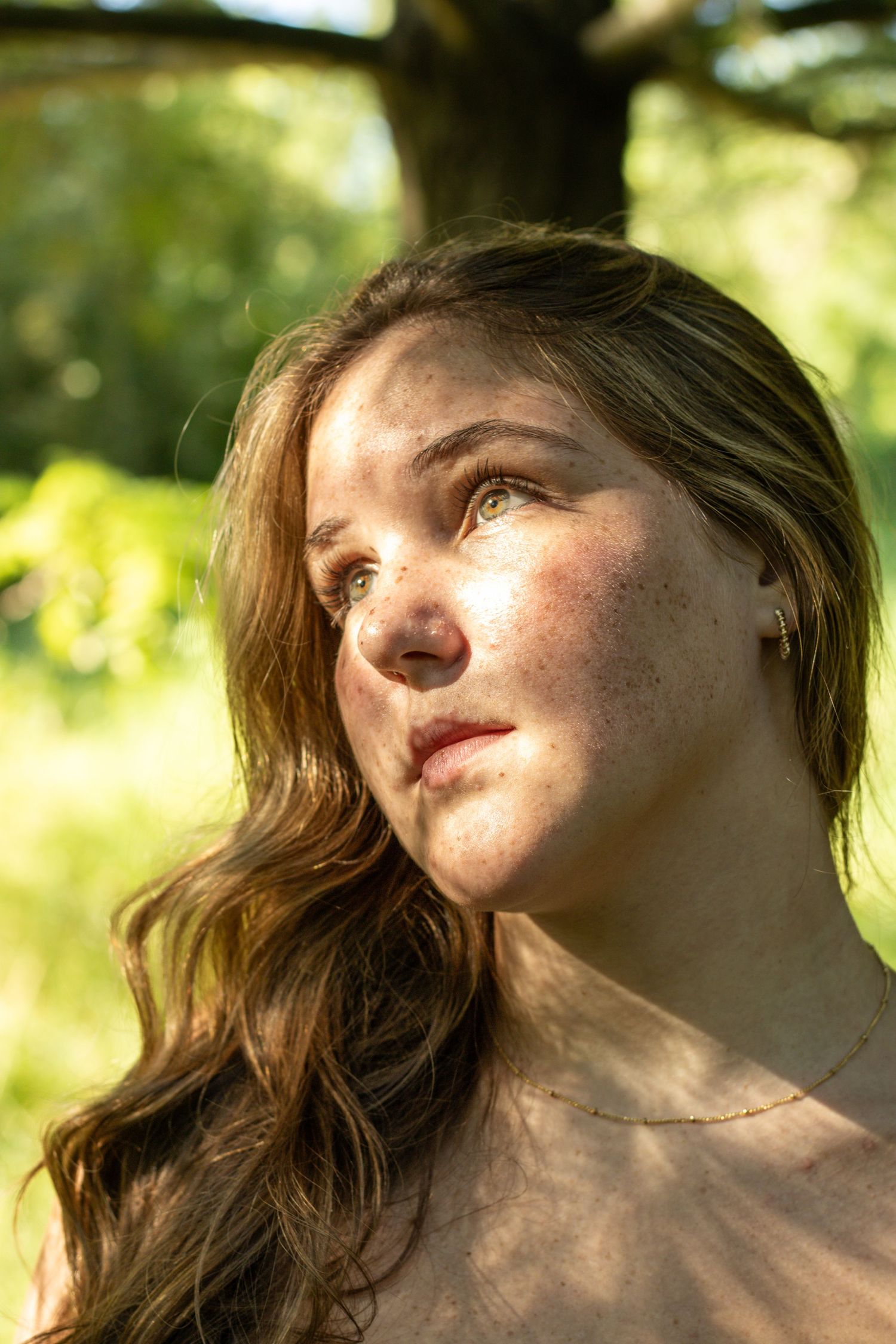 Young woman with freckles and wavy brown hair gazing upward in dappled forest sunlight.