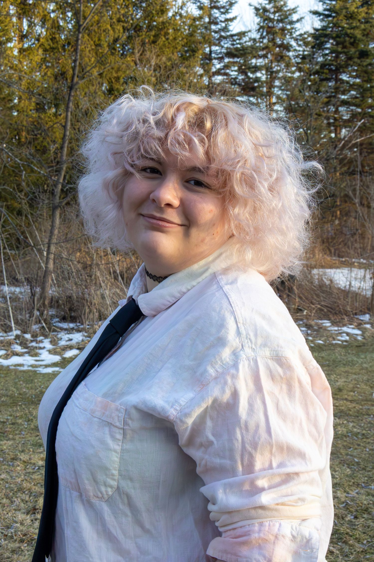Woman with curly pink-blonde hair smiling outdoors in a light pink jacket near snowy trees.
