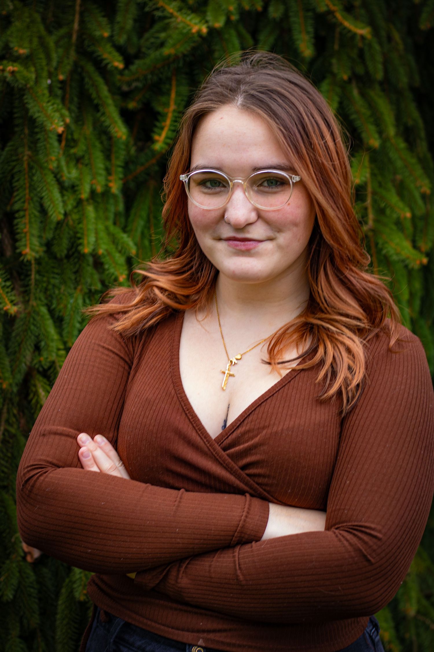 Young woman with red hair and clear glasses wearing a brown wrap top with arms crossed in front of green evergreen trees.