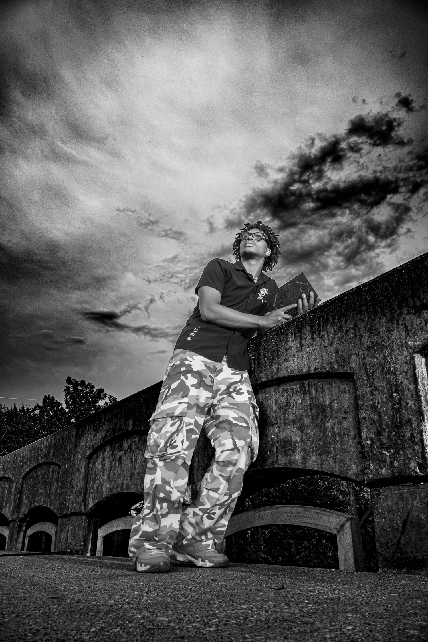 Black and white photo of a person in camo pants leaning against a stone wall under a dramatic cloudy sky.