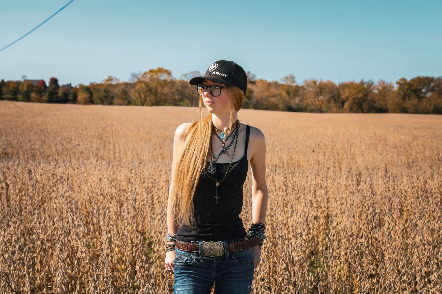 Young woman with long blonde hair wearing a black cap and tank top standing in a golden field under blue sky.
