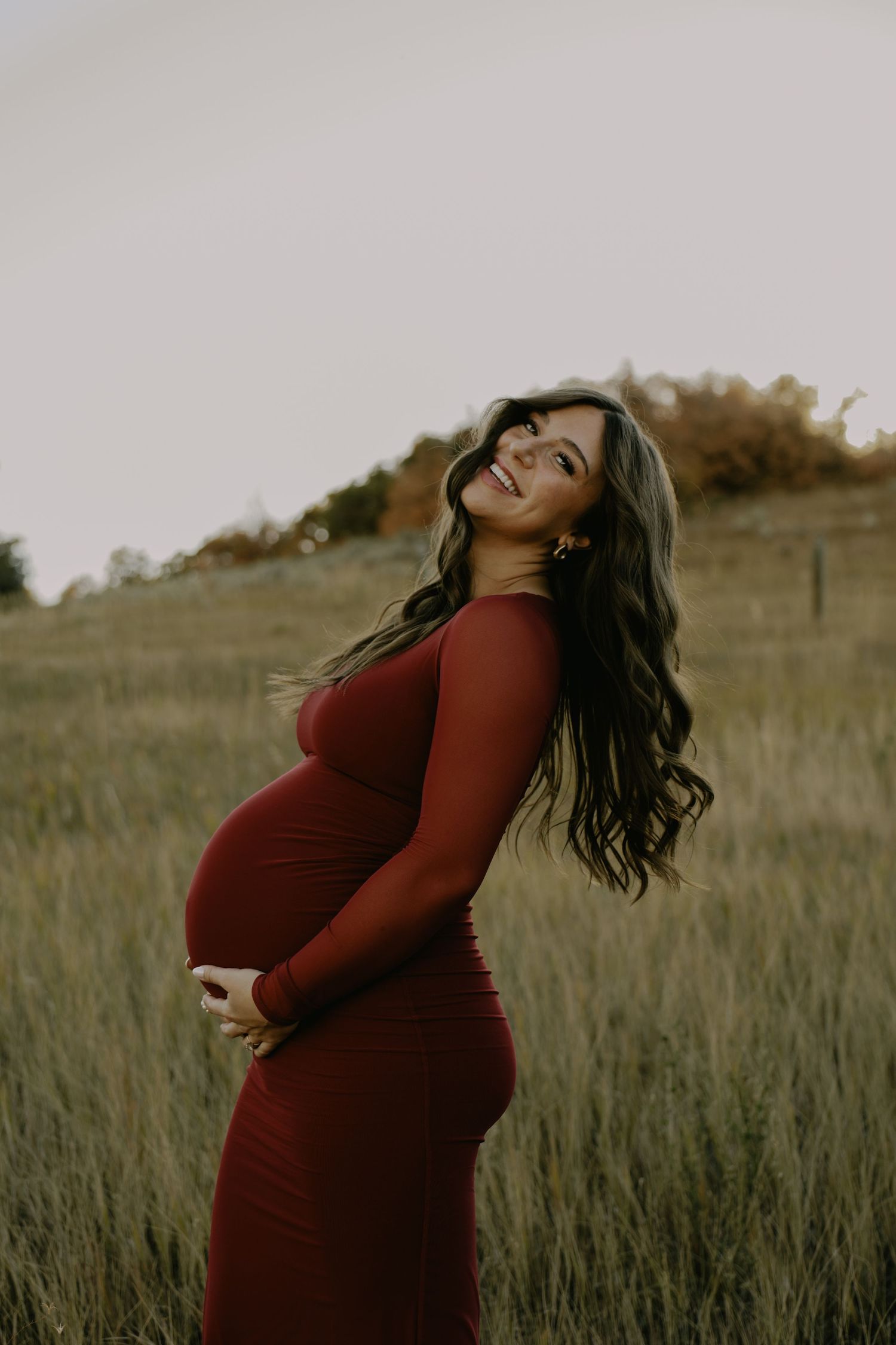 Expectant mother in red dress stands joyfully in tall grass field.