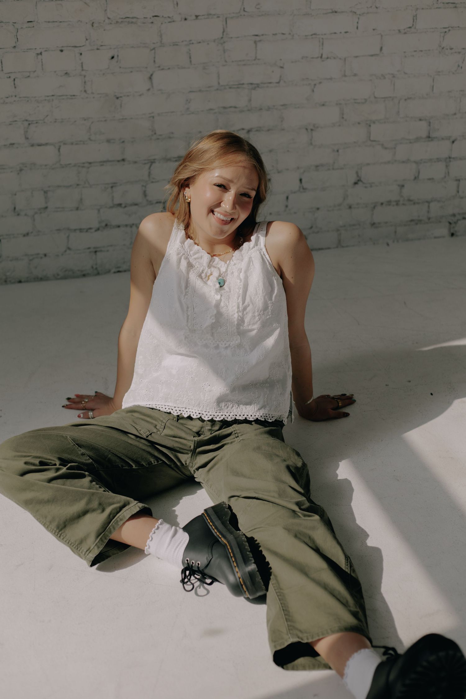 Person in white lace top and olive pants lounging on white floor with dramatic lighting.