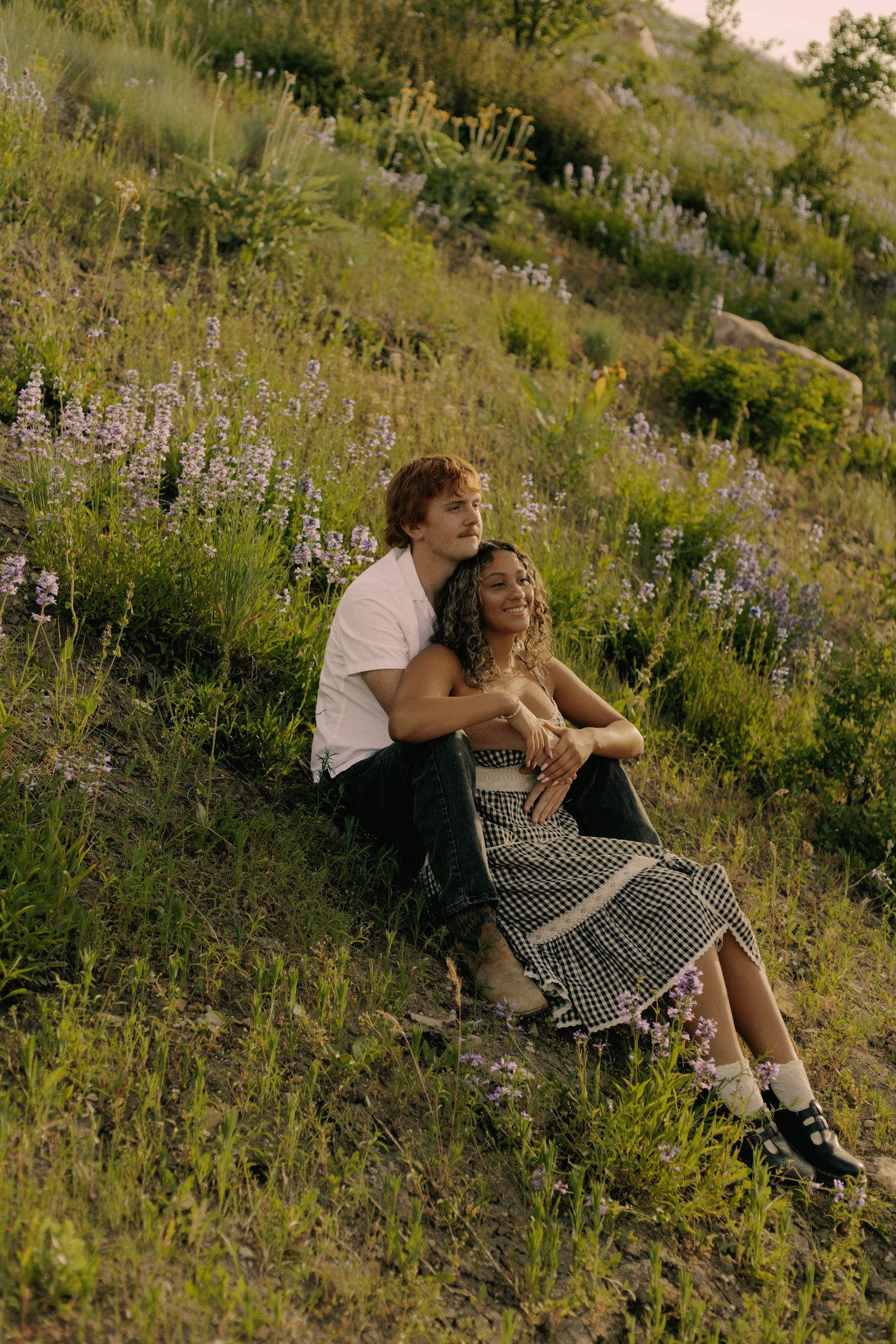 Couple sitting close together on hillside covered in purple wildflowers.
