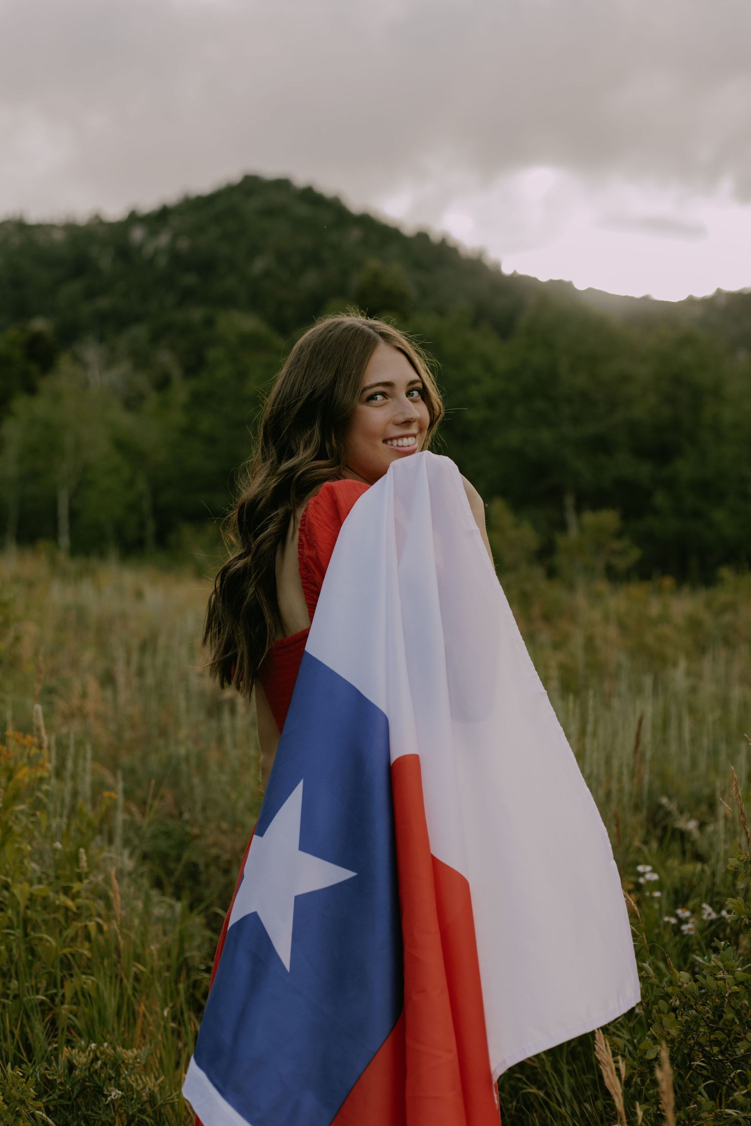 Someone wrapped in a Texas state flag stands in a meadow with mountains in the background.