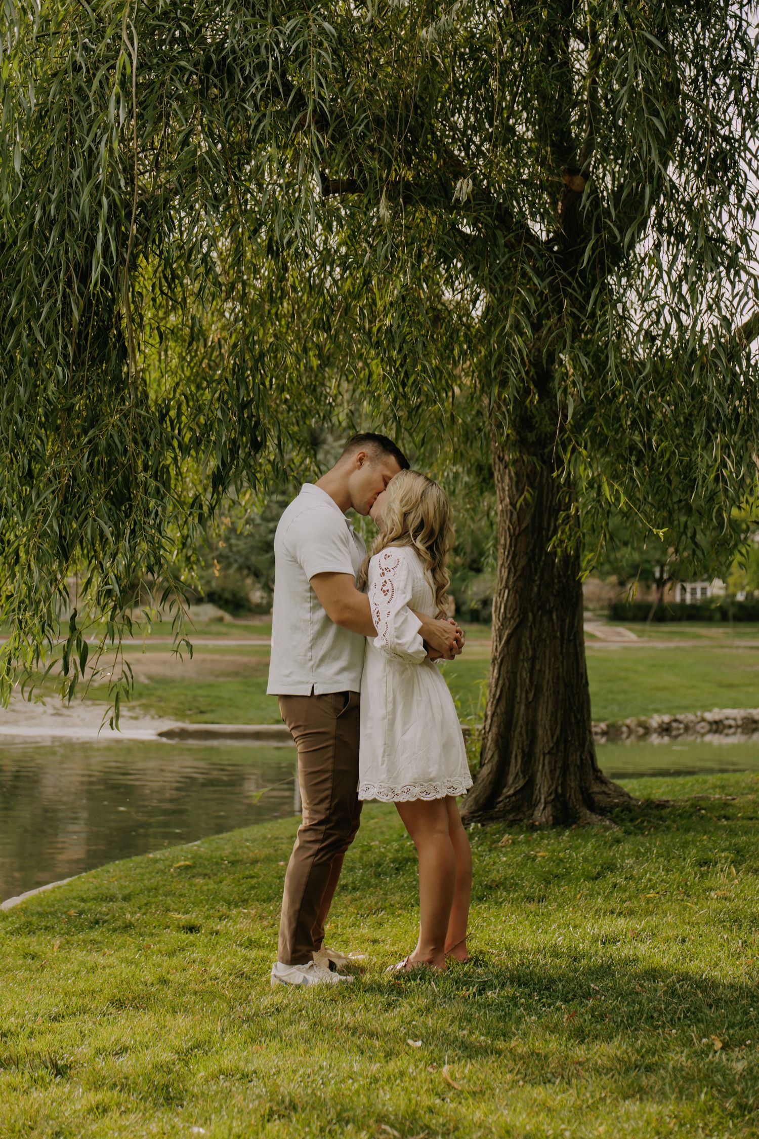 Intimate couple sharing a tender moment beneath drooping willow branches by a pond.