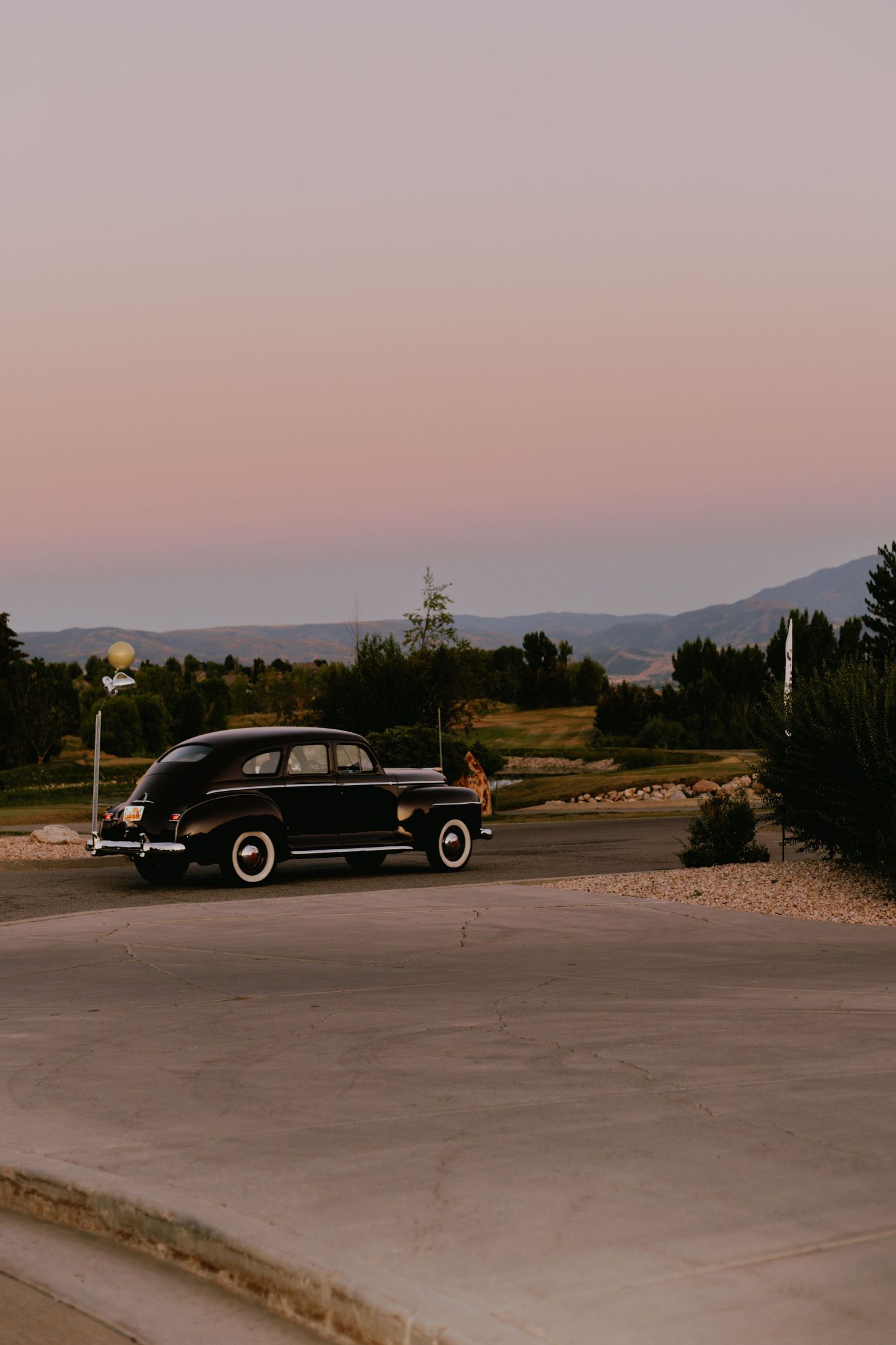 Vintage black car parked in an empty lot with mountains visible in the background at sunset.