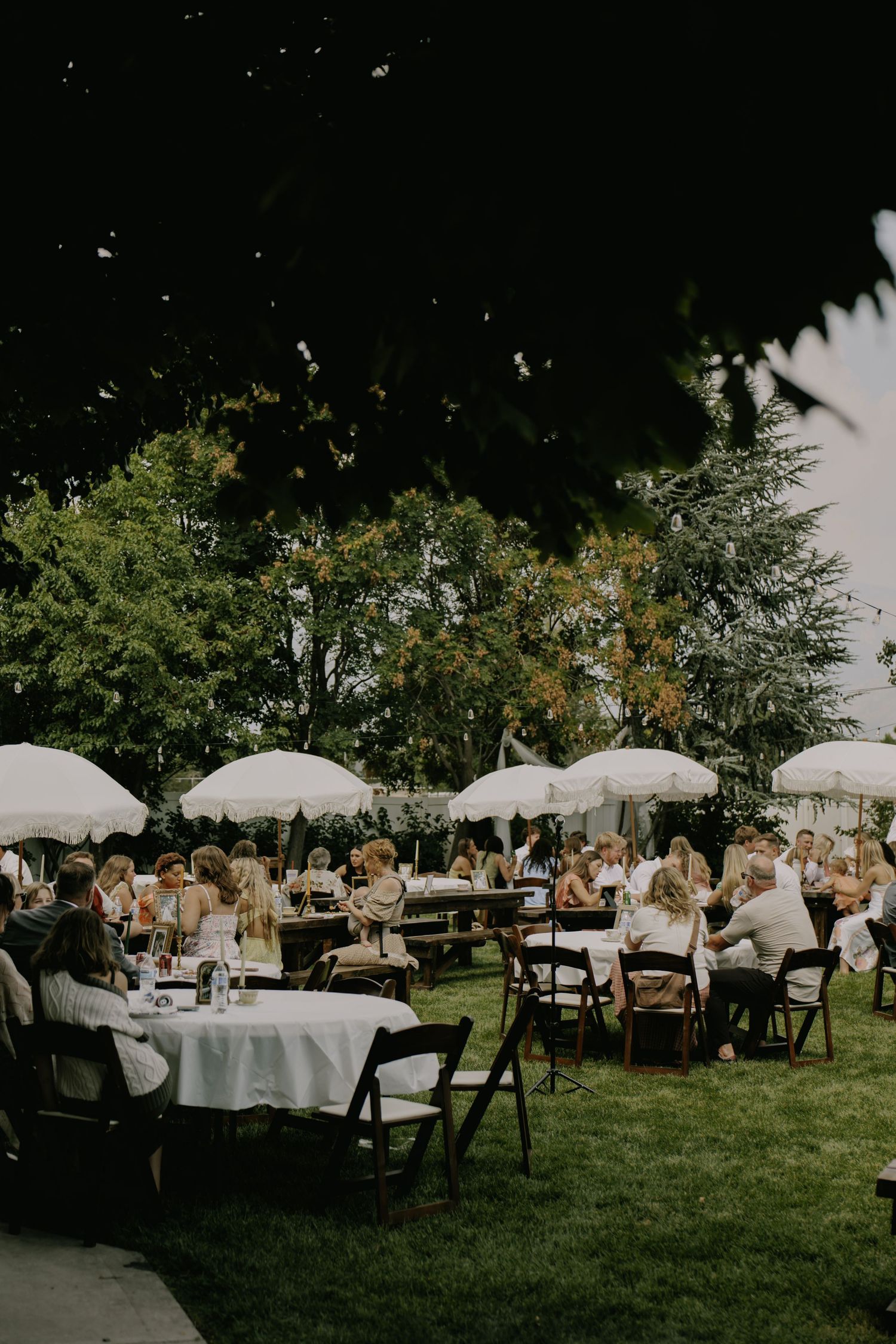 Outdoor wedding reception with white umbrellas and round tables on a sunny lawn.