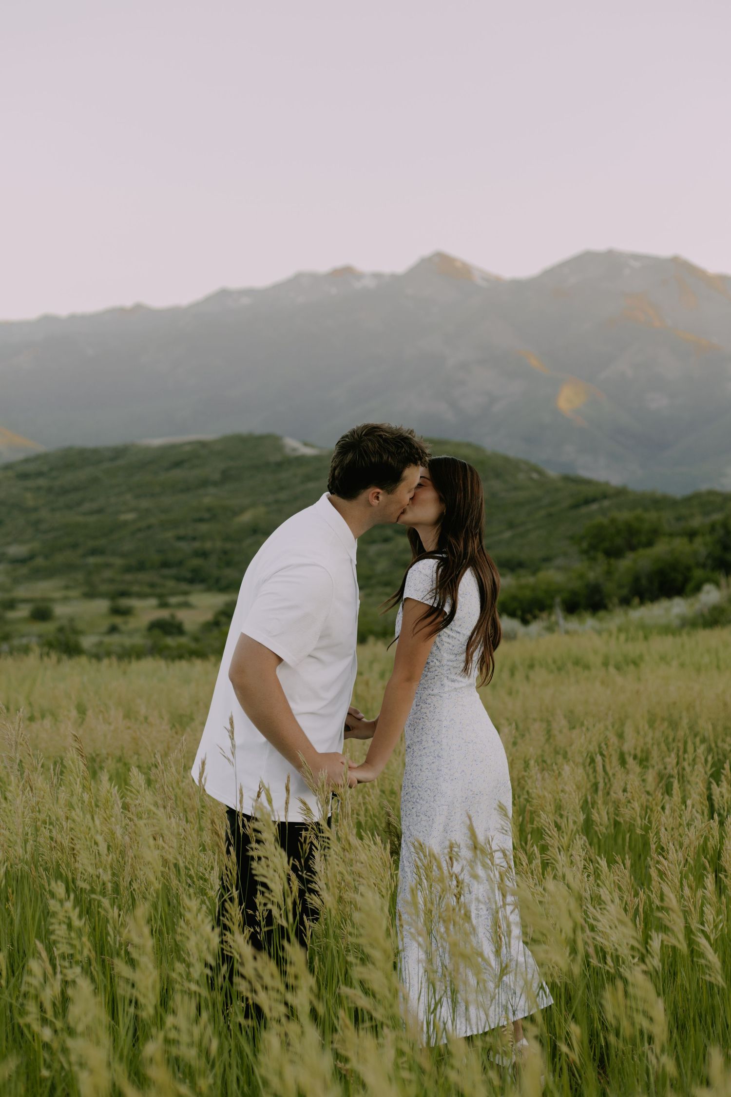 Romantic moment shared between couple in white clothing standing in tall grass with mountains behind.