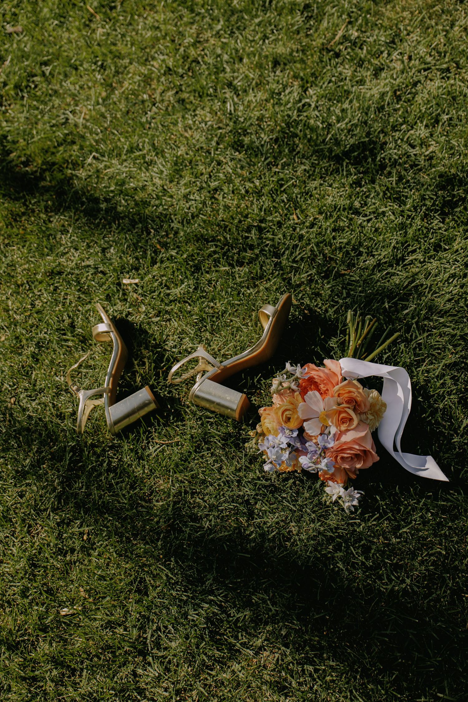 Wedding shoes and peach floral bouquet arranged on green grass in natural lighting.