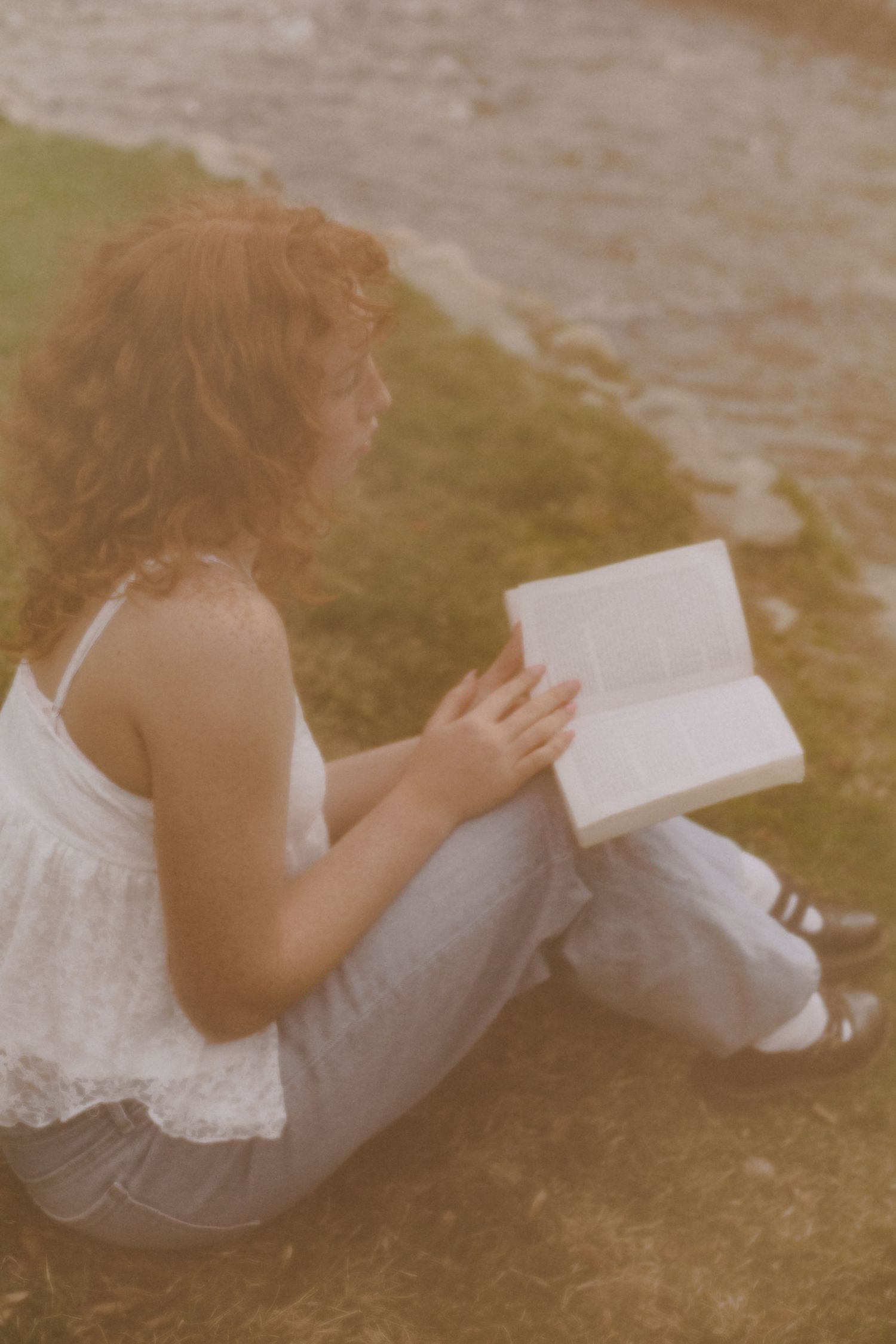 Person in white dress reading a book while sitting on grass near water.