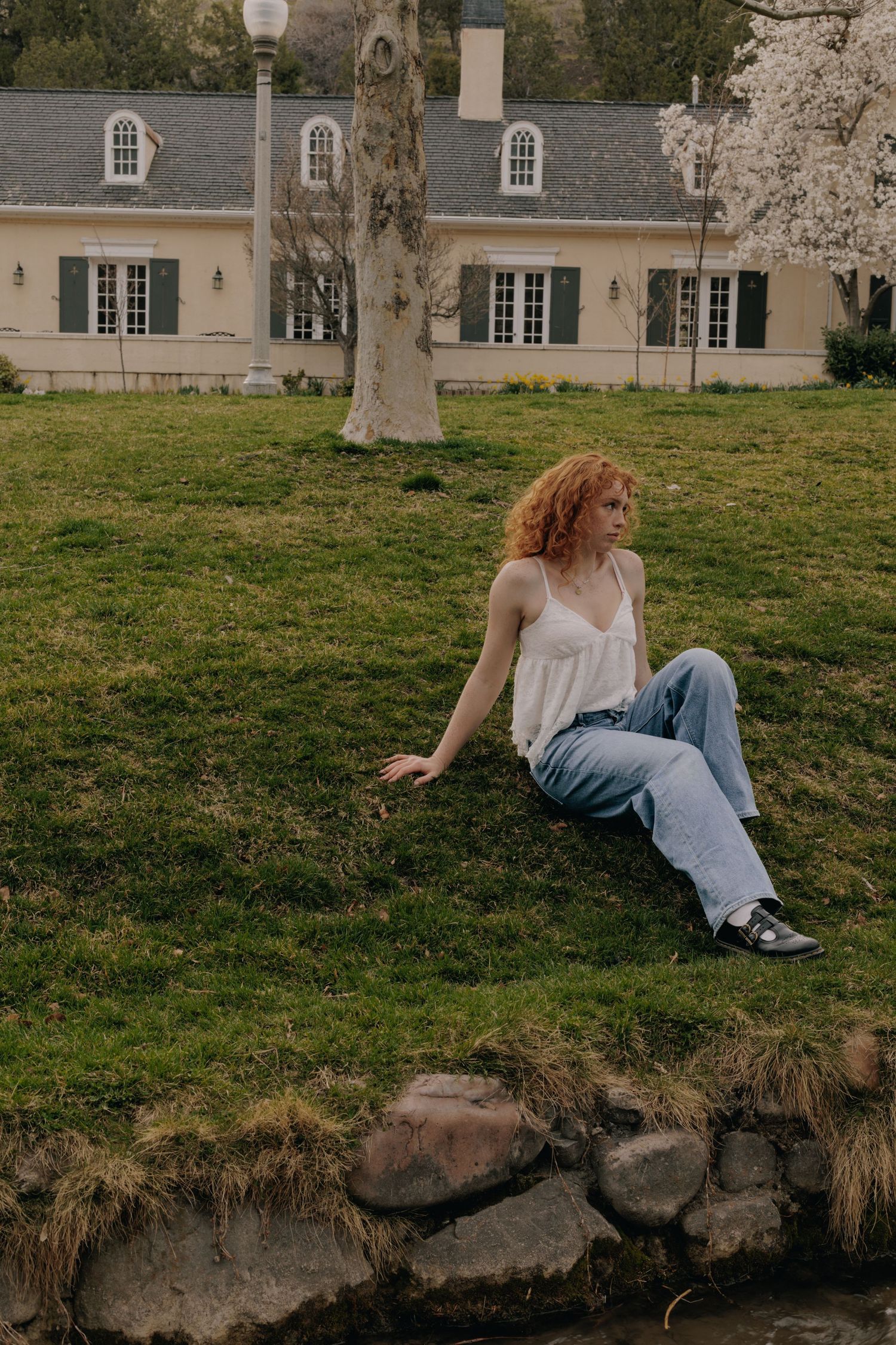Someone in white lounges by a creek bank with historic buildings in the background.