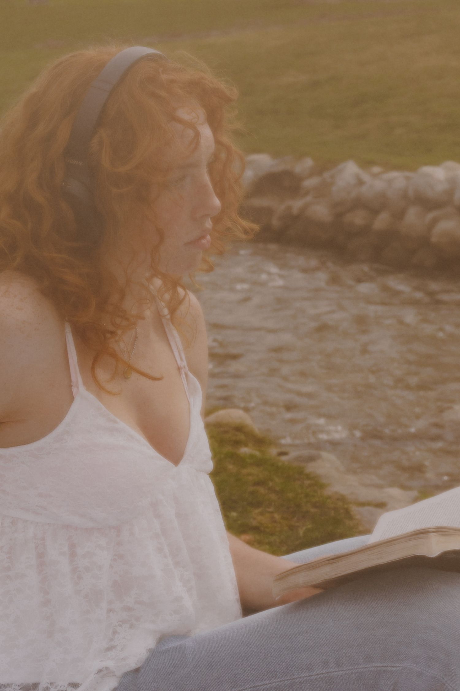Person in white dress reading by waterside in warm sunlight.