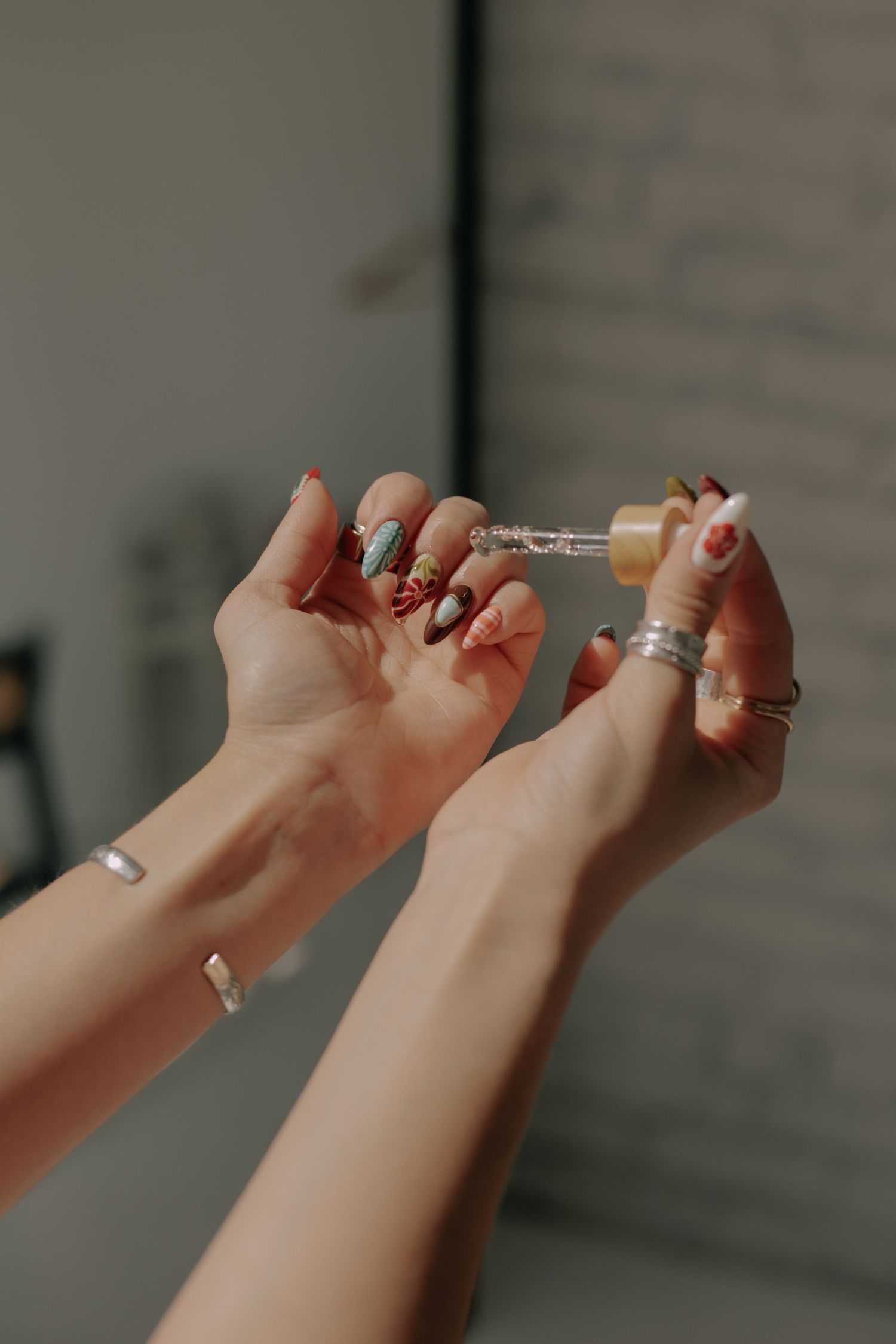 Hands with painted nails and rings holding manicure tools against a light background.