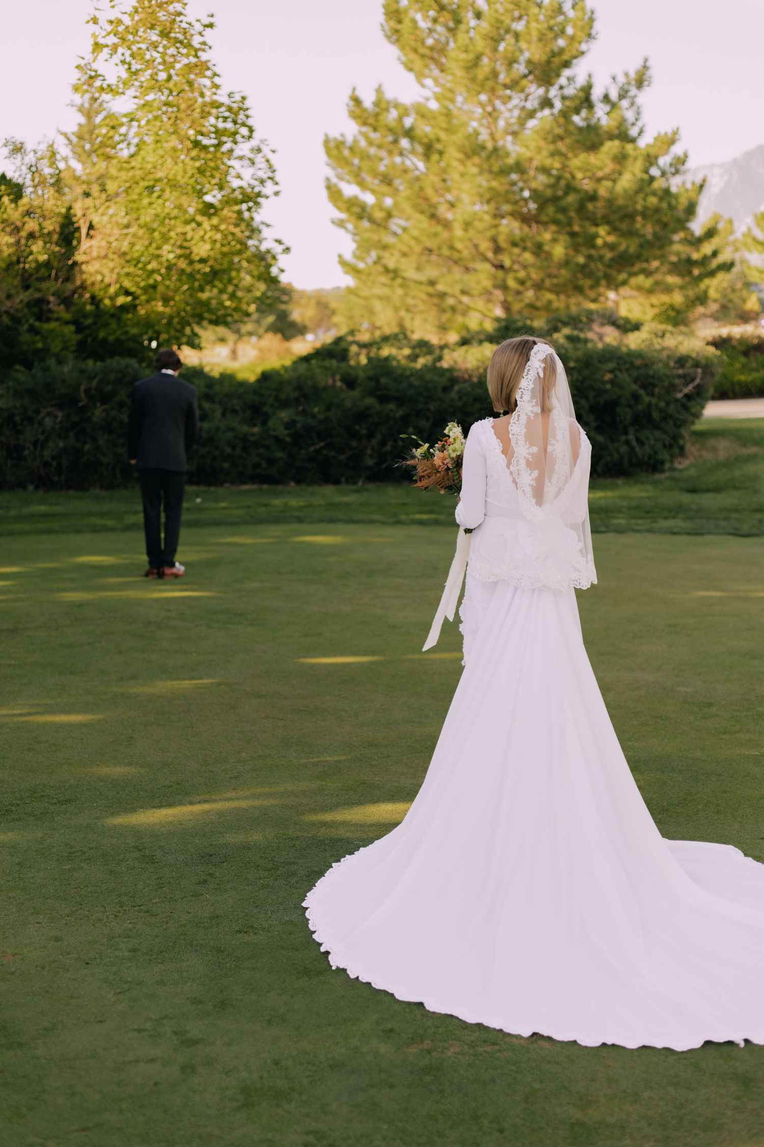 Wedding dress train flowing across green lawn with trees in background during golden hour.