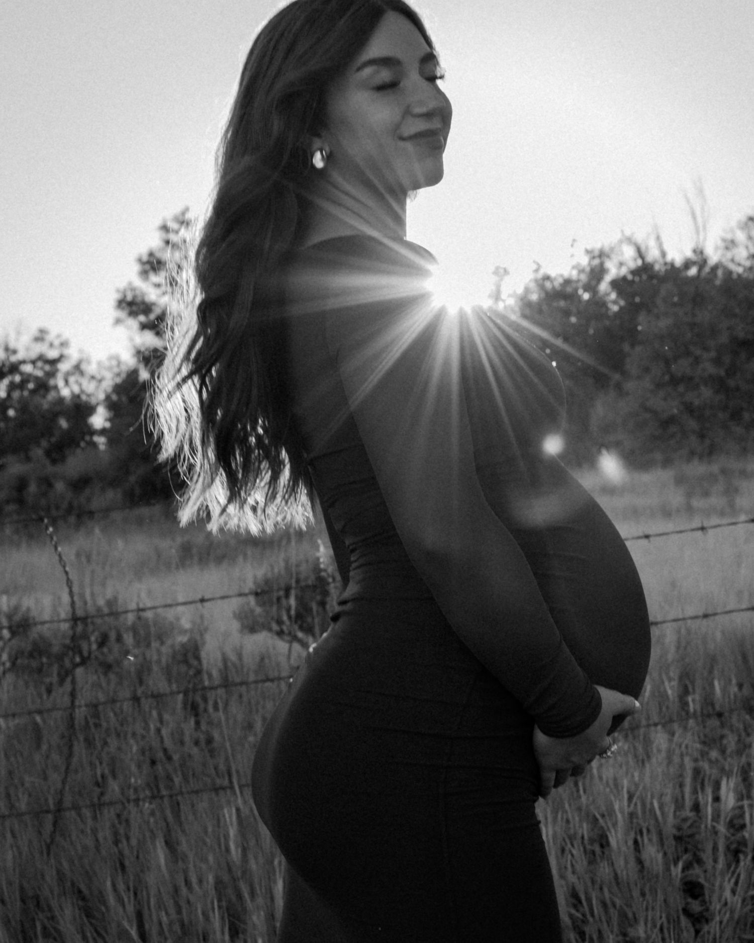 Black and white silhouette of pregnant woman in field with dramatic sun flare.