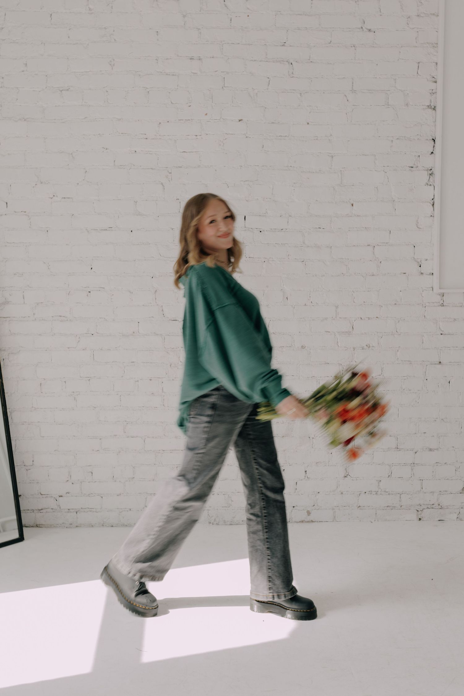 Person in green sweater and gray pants dancing playfully against white backdrop.