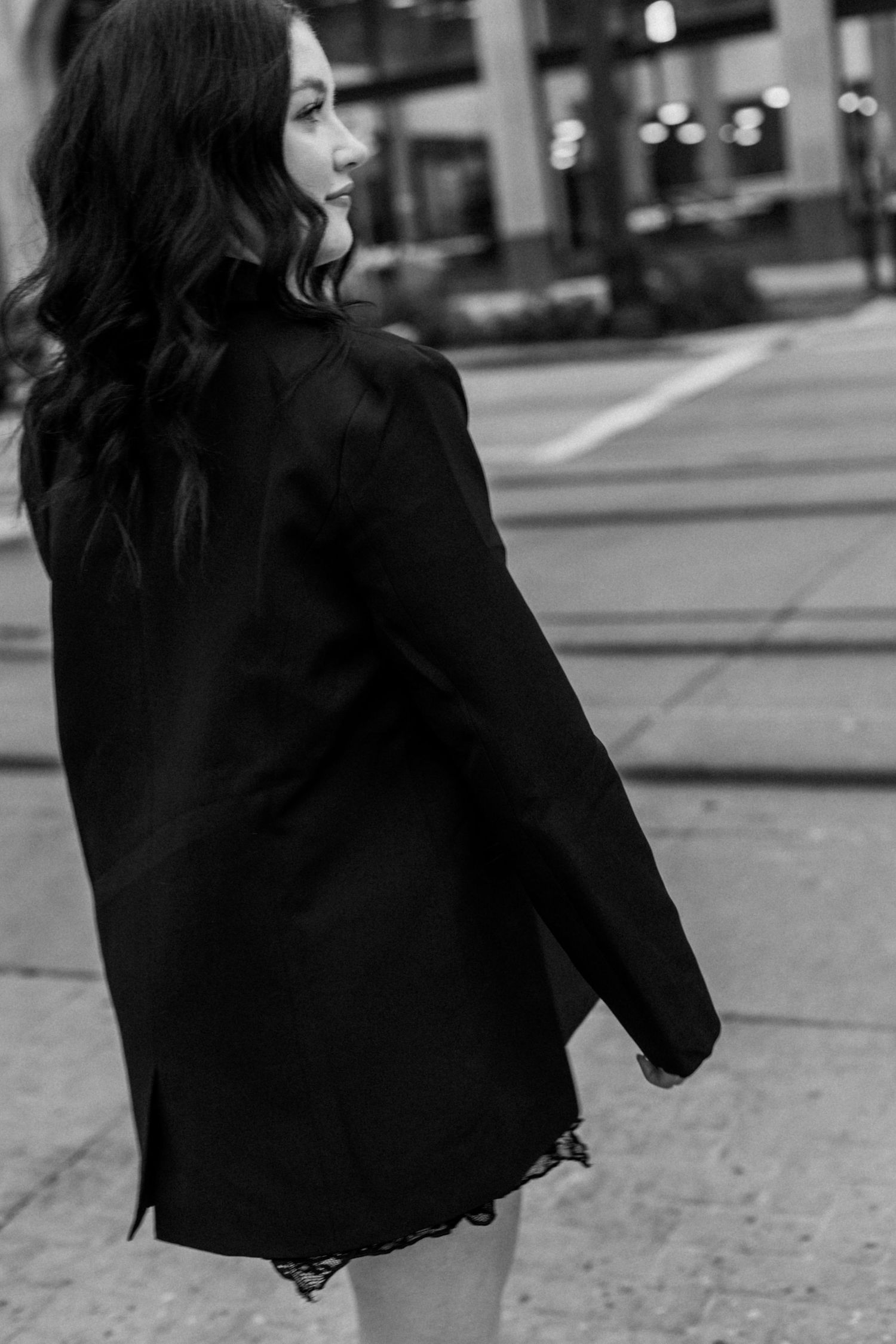 A person wearing a black jacket walks along a city street in a stylish urban setting.