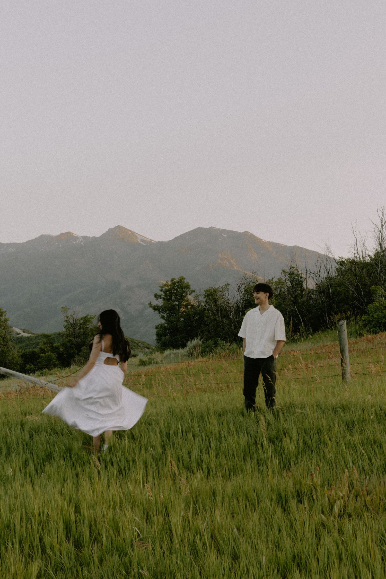 A couple dances playfully in a grassy field with mountains in the background at sunset.