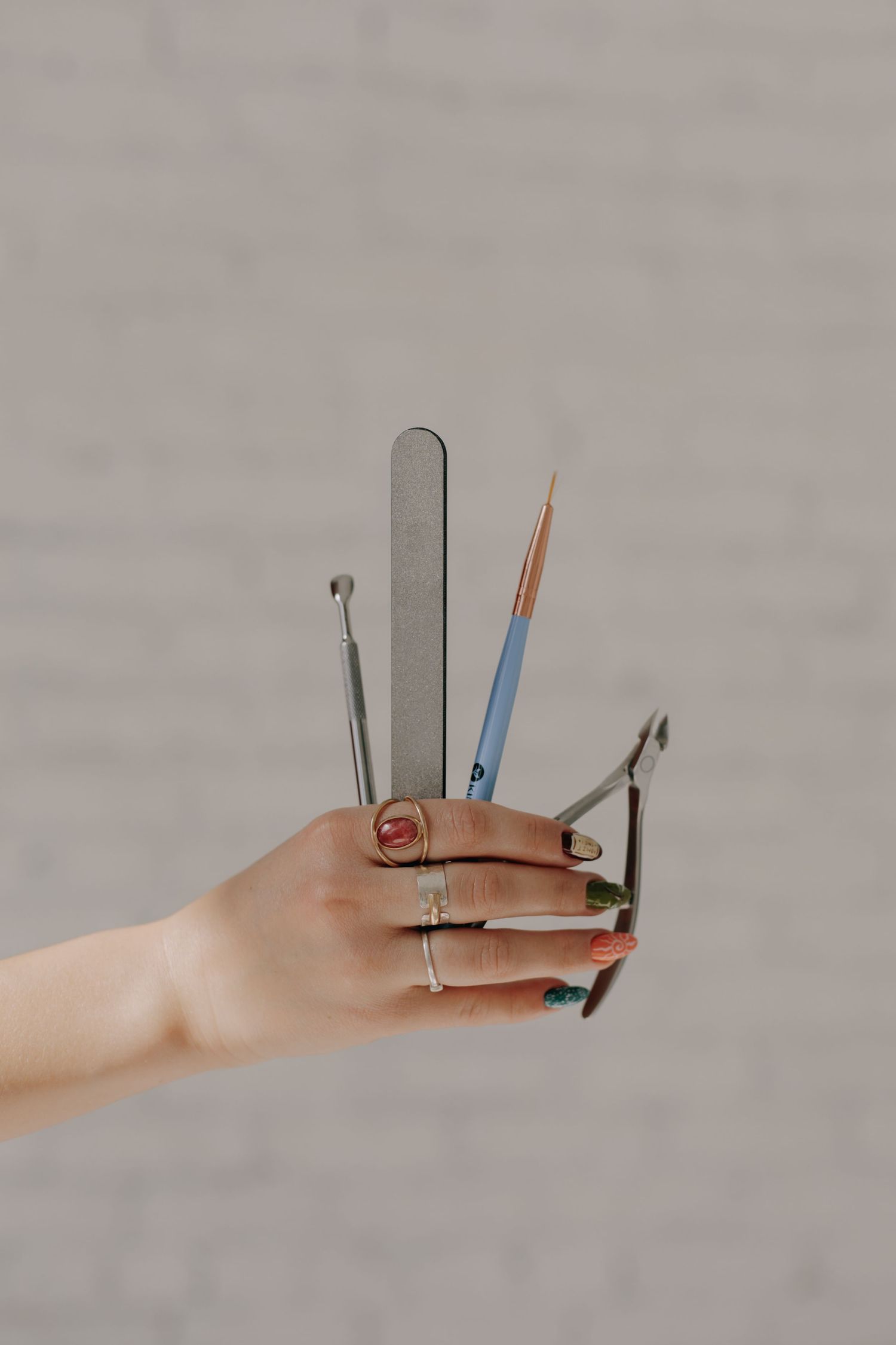 Hand displaying various manicure tools including nail file and clippers against gray background.