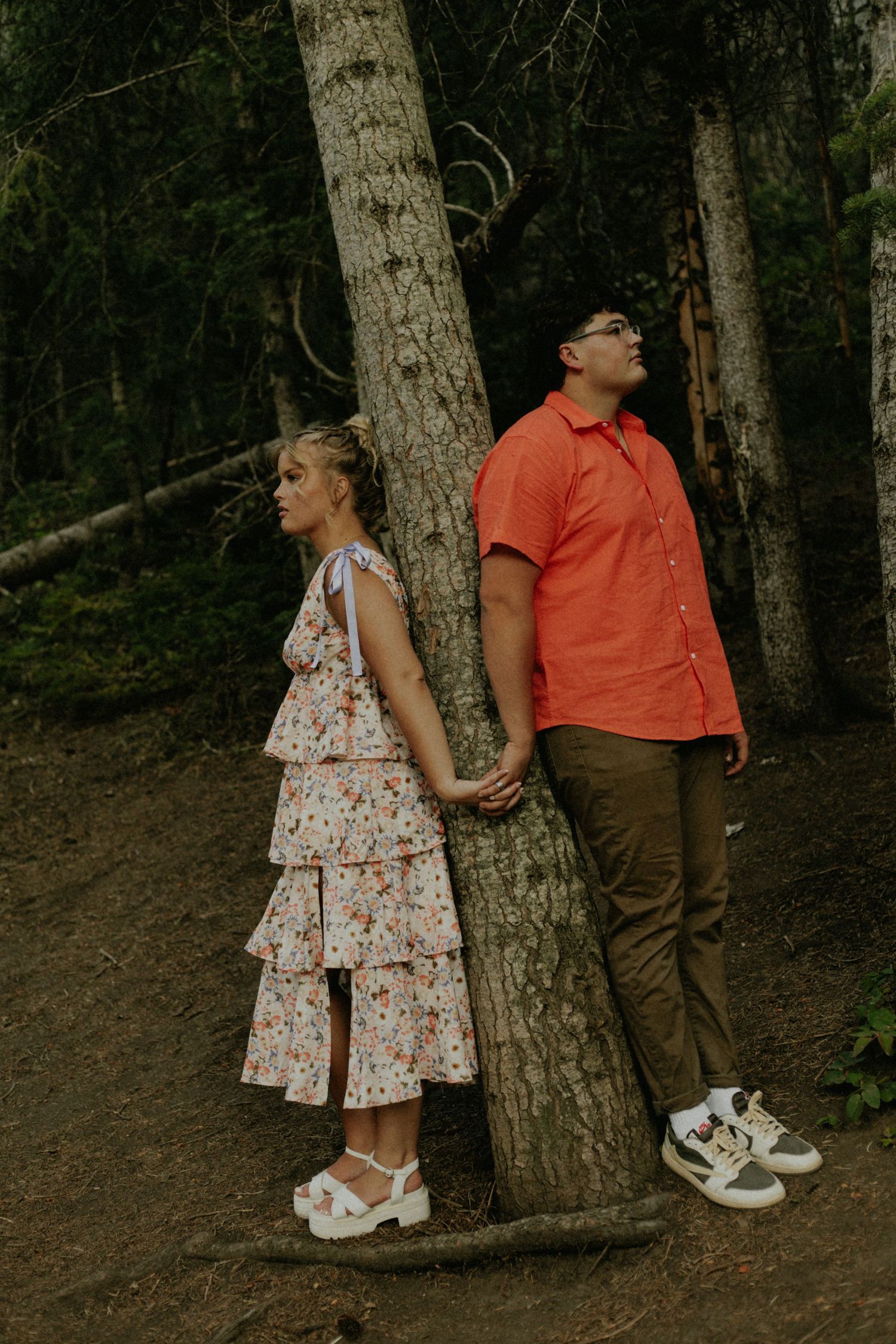 A playful couple poses by tree trunks in a forest setting wearing contrasting outfits.