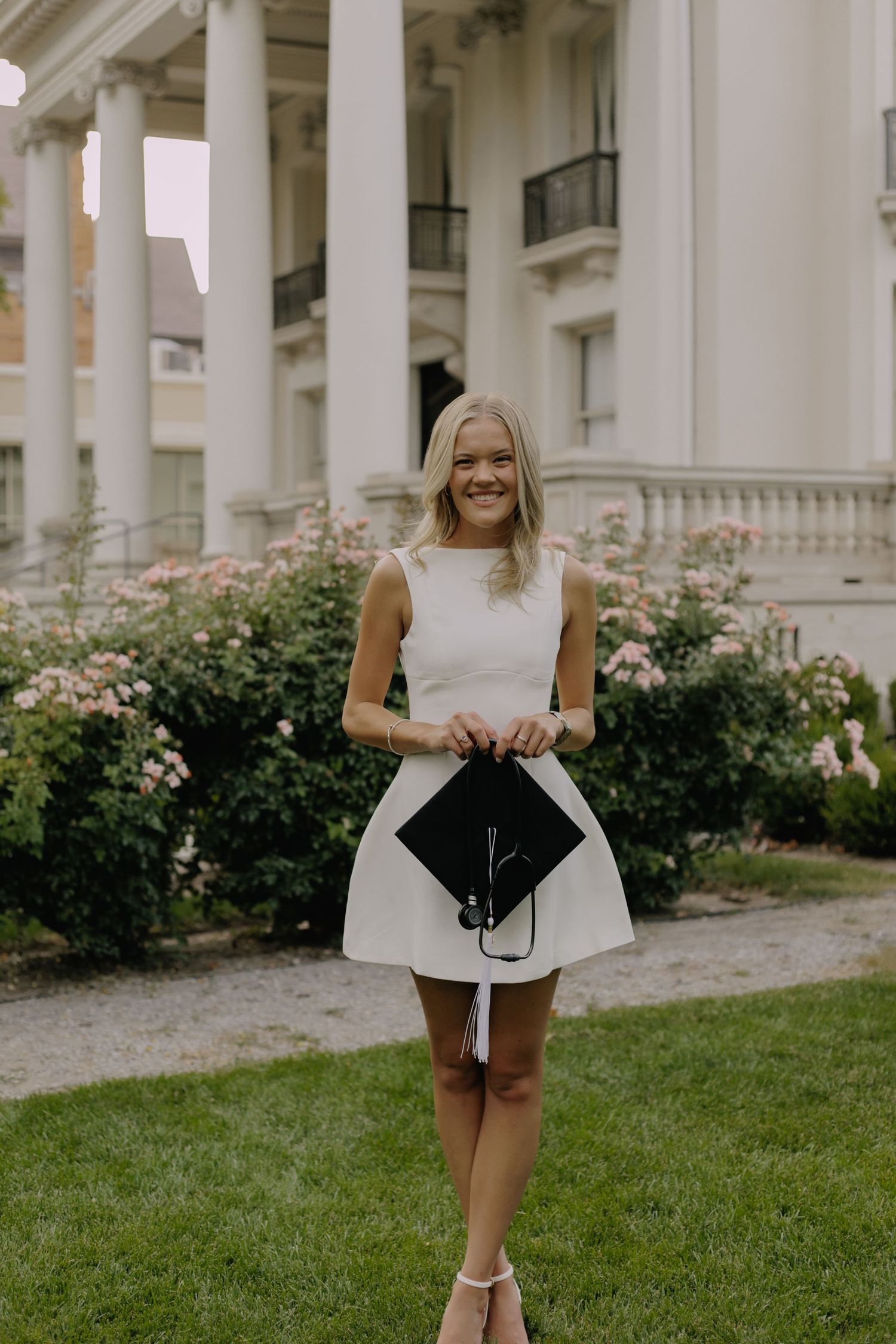 Graduate in white dress stands on grass near ornate building with white columns.