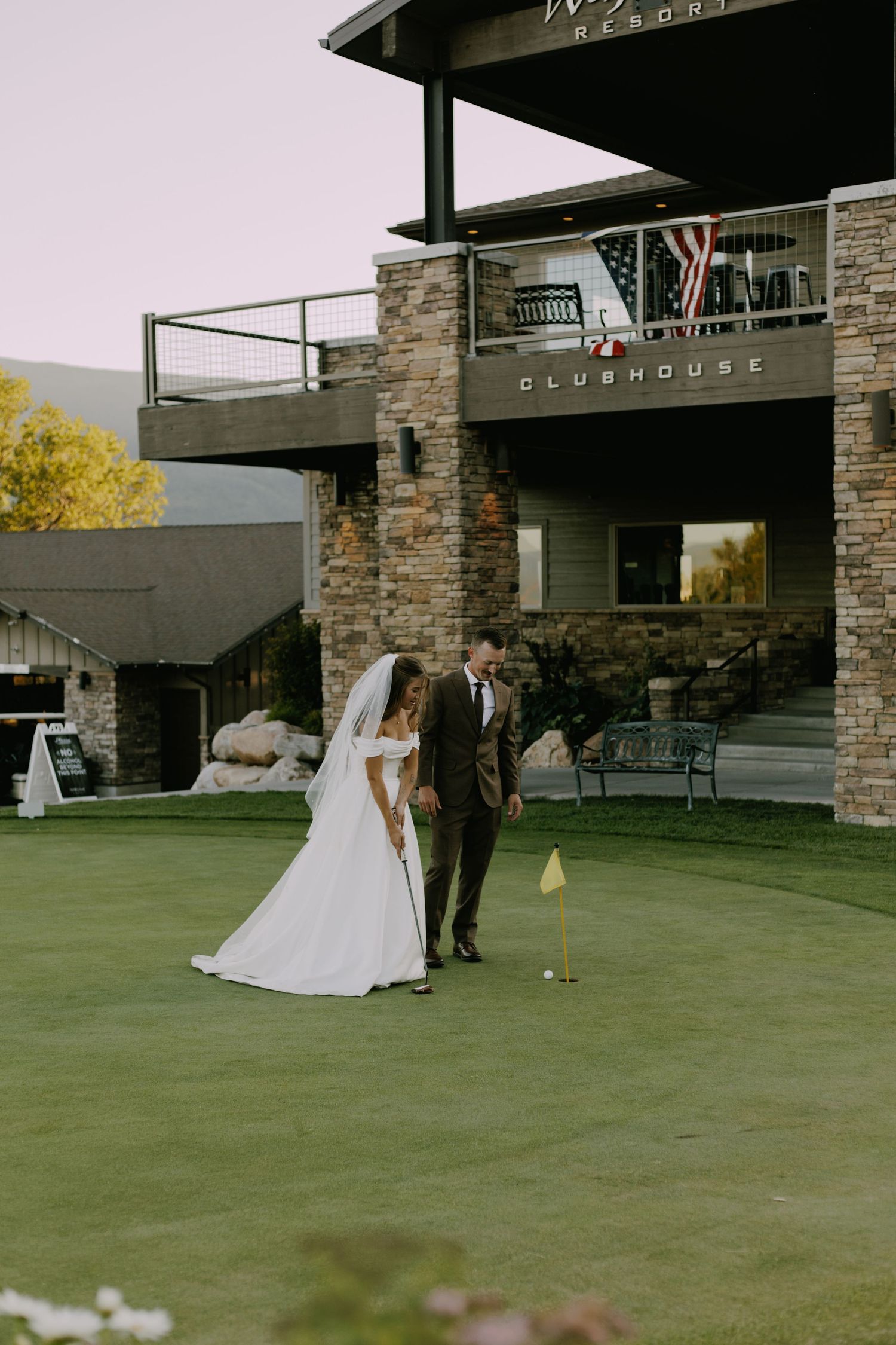 A wedding party plays lawn games outside a modern stone clubhouse building on a sunny day.