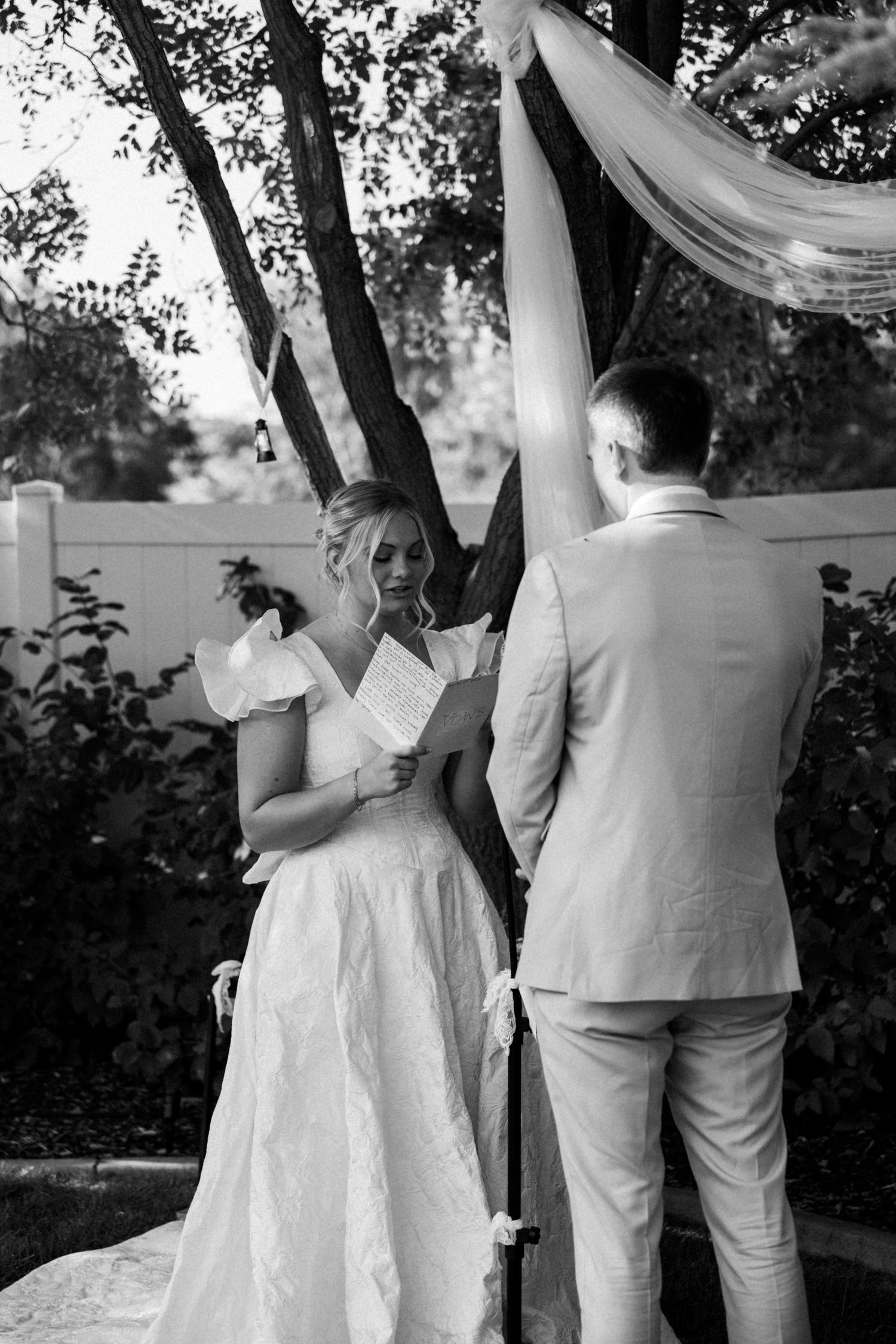 A couple in white attire dance together under trees at an outdoor wedding celebration.