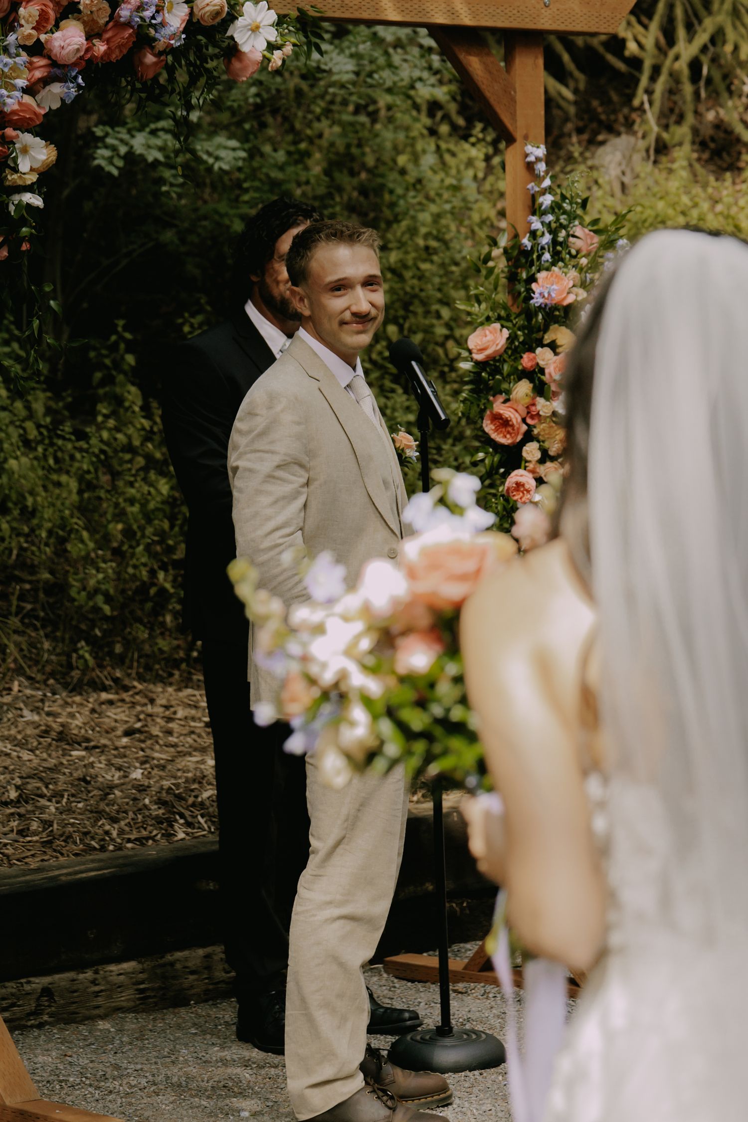 Wedding ceremony scene with floral arch decorations and guests seated outdoors.
