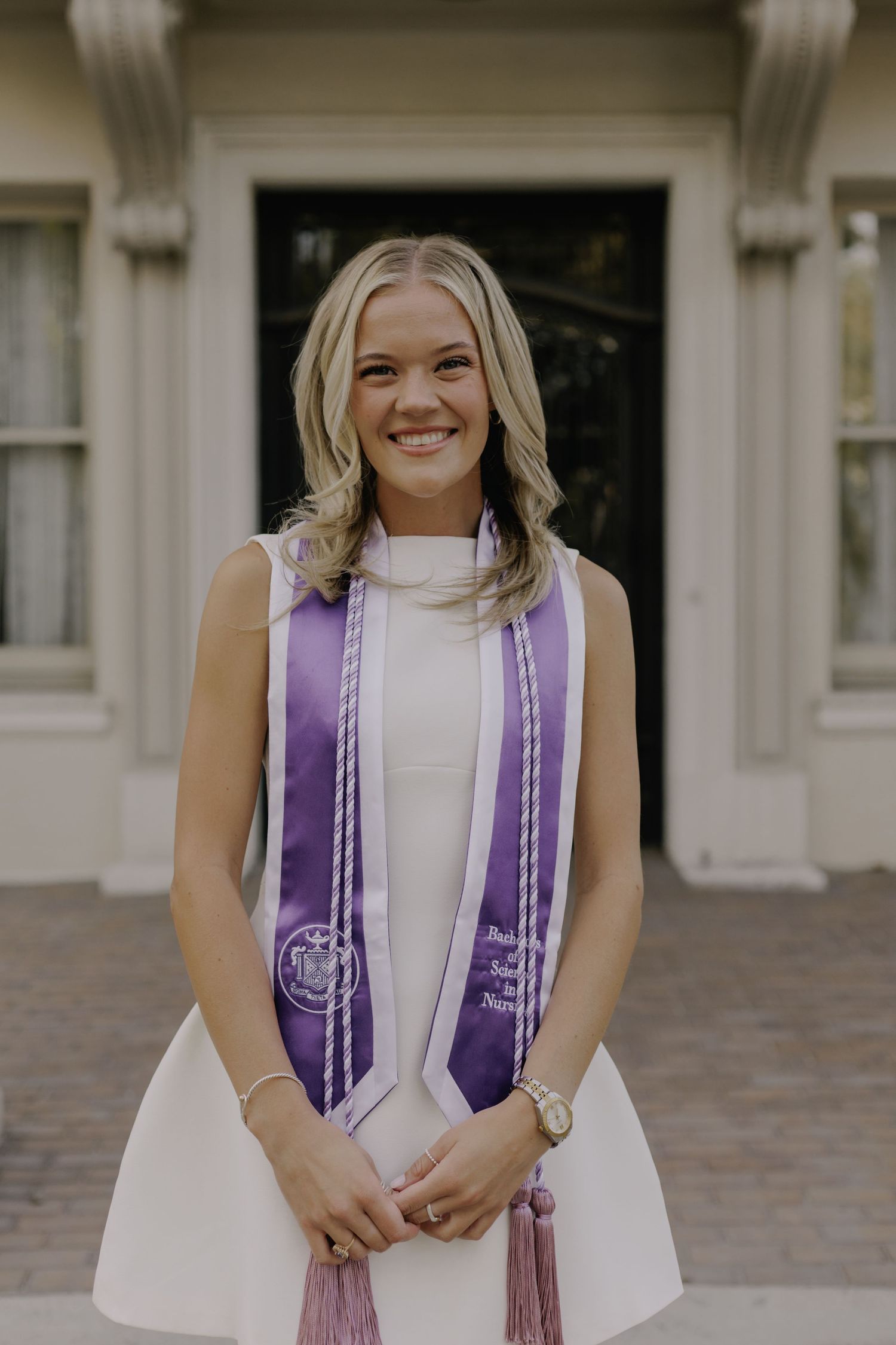 Graduate wearing purple and white graduation stoles smiling in front of building columns.