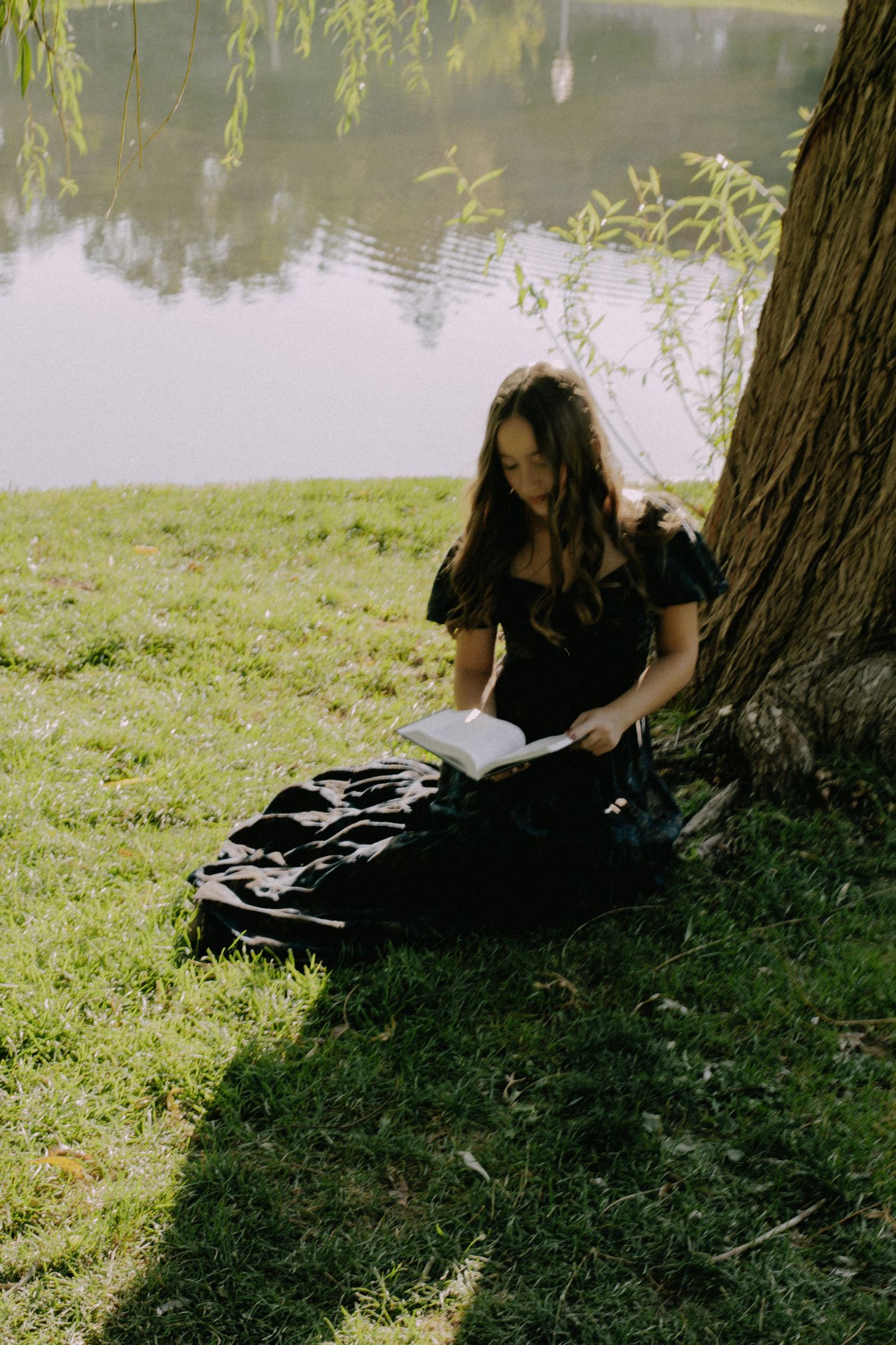 Figure in dark dress relaxes under willow tree by peaceful pond.
