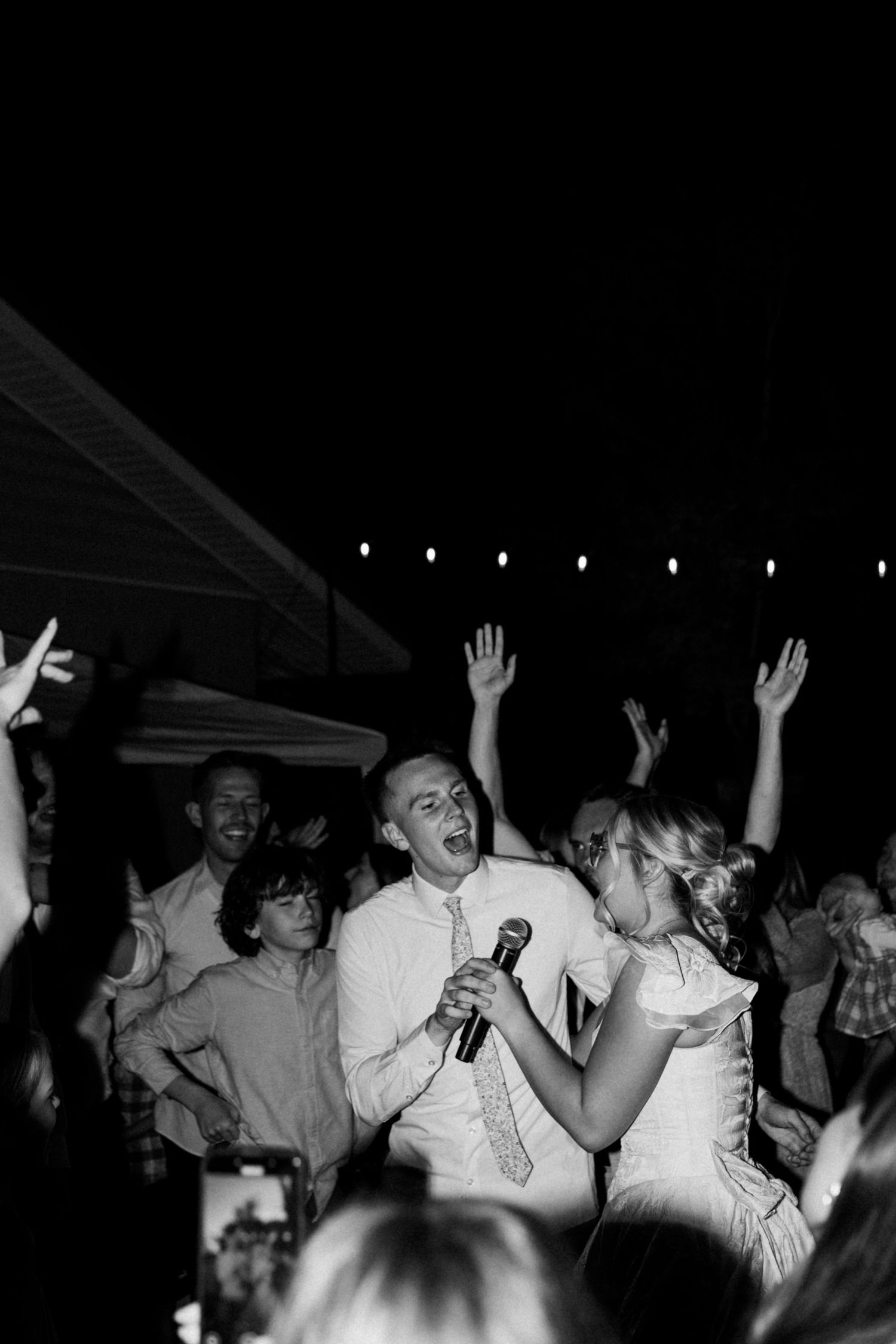 Joyful wedding reception crowd dancing and singing with hands in the air at night.