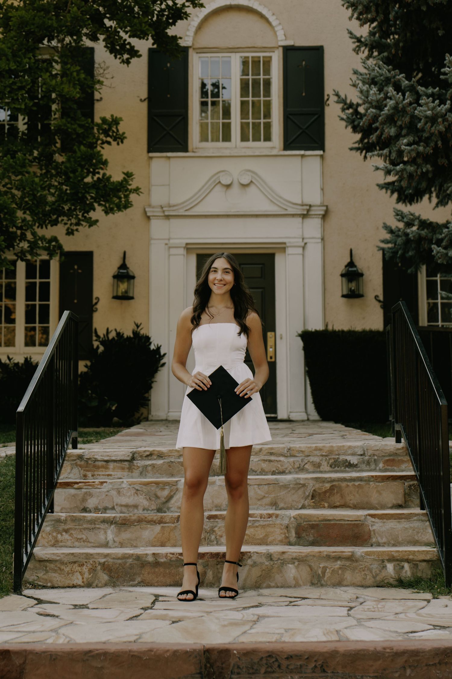 Person in graduation attire poses on architectural stone steps of building.