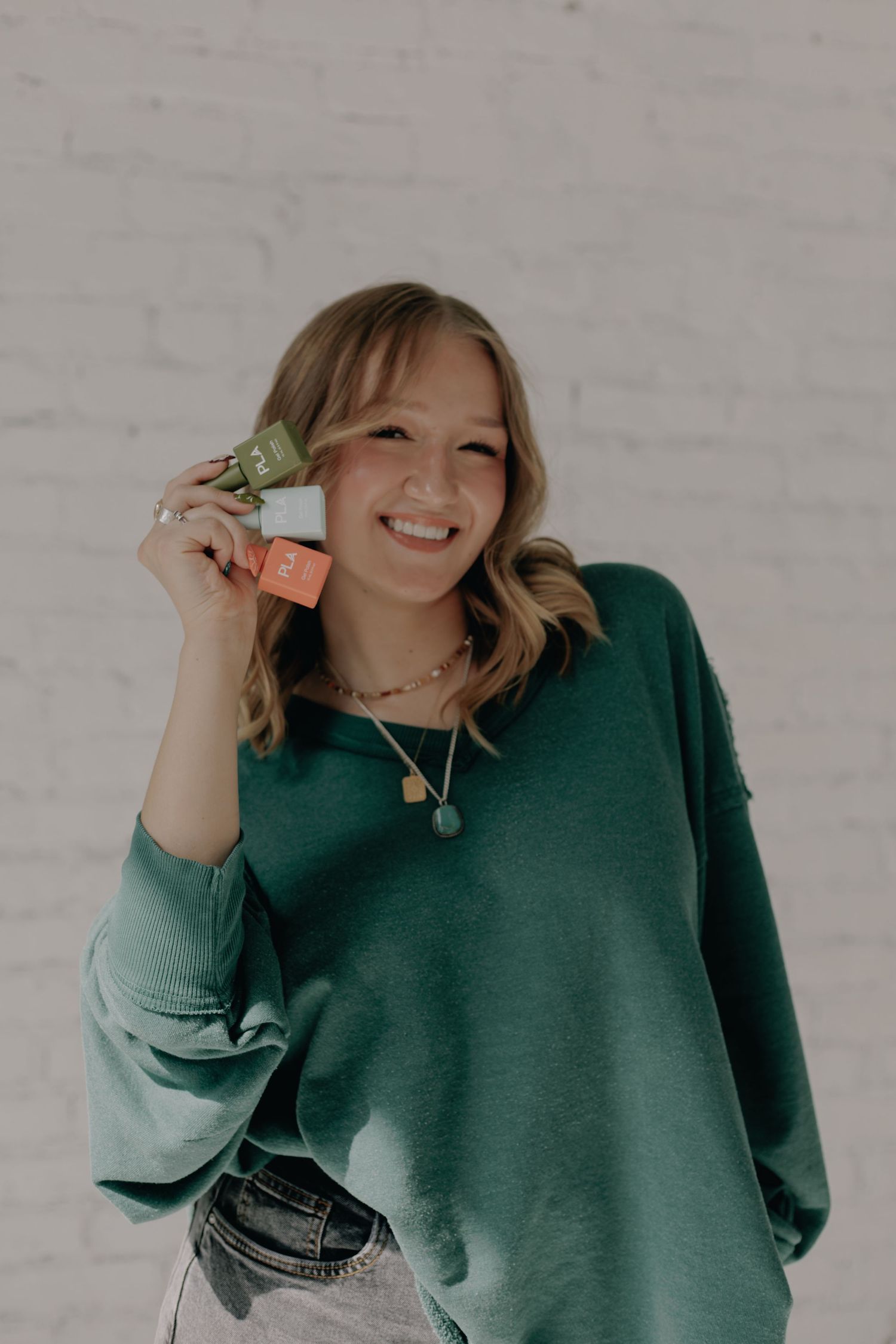 Person in green sweater playfully poses against white brick wall holding small colorful bottles.