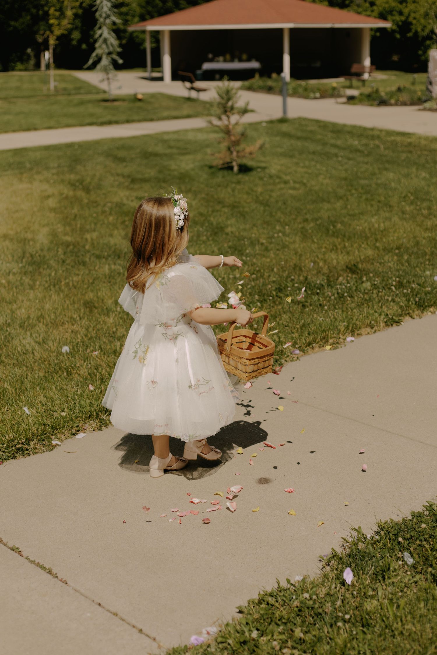 Young flower girl in white dress scattering rose petals along a garden pathway.